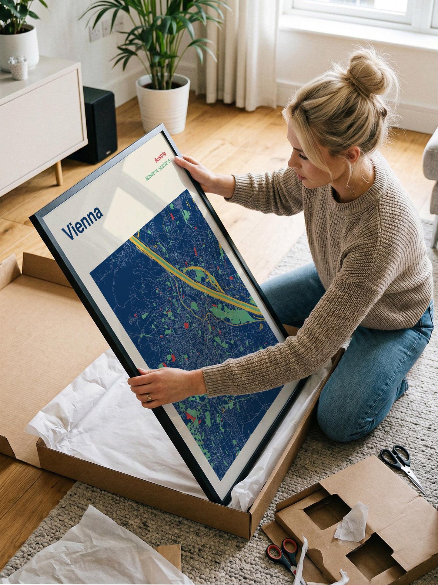 A woman is kneeling on the floor and holding a framed map of Vienna, with a potted plant and other items visible in the background.