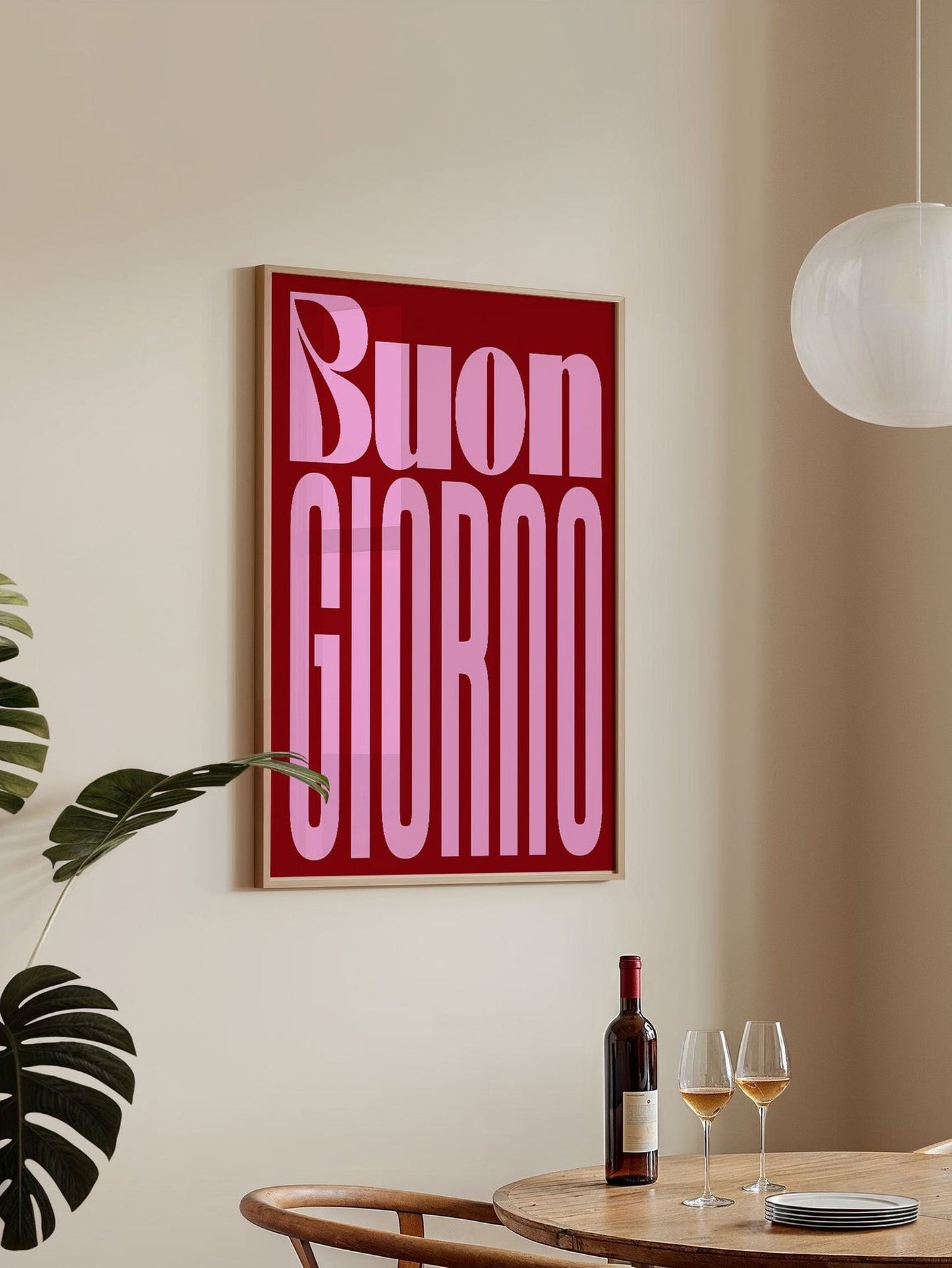 a framed poster with the text "Buon Giordano" in a pink color, hanging on a wall above a dining table with two wine glasses and a bottle of wine.