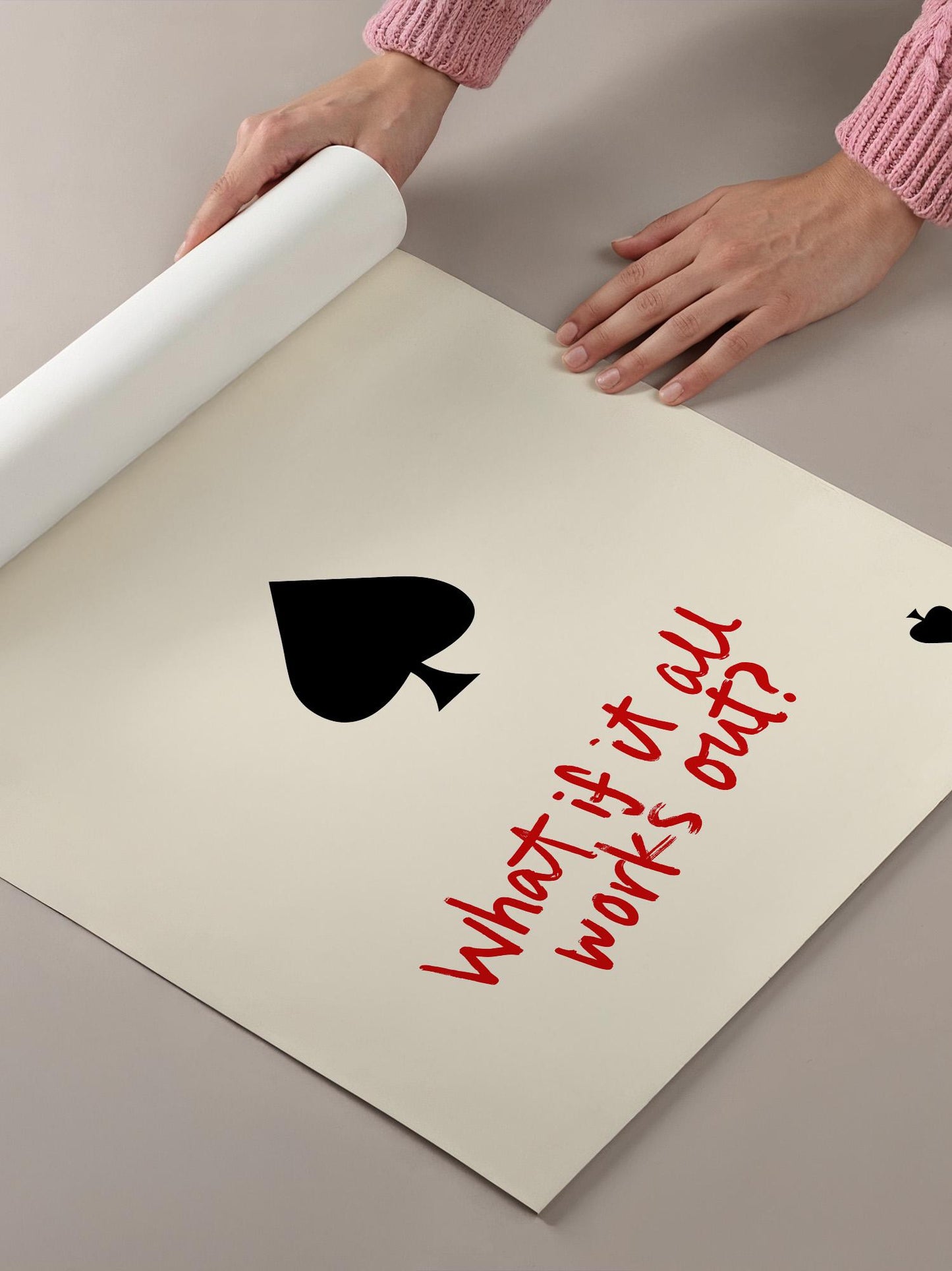 A person's hands are holding a roll of paper, which is laid out on a table. The paper has a black symbol resembling a spade and the text "What if it all works out?" written on it.