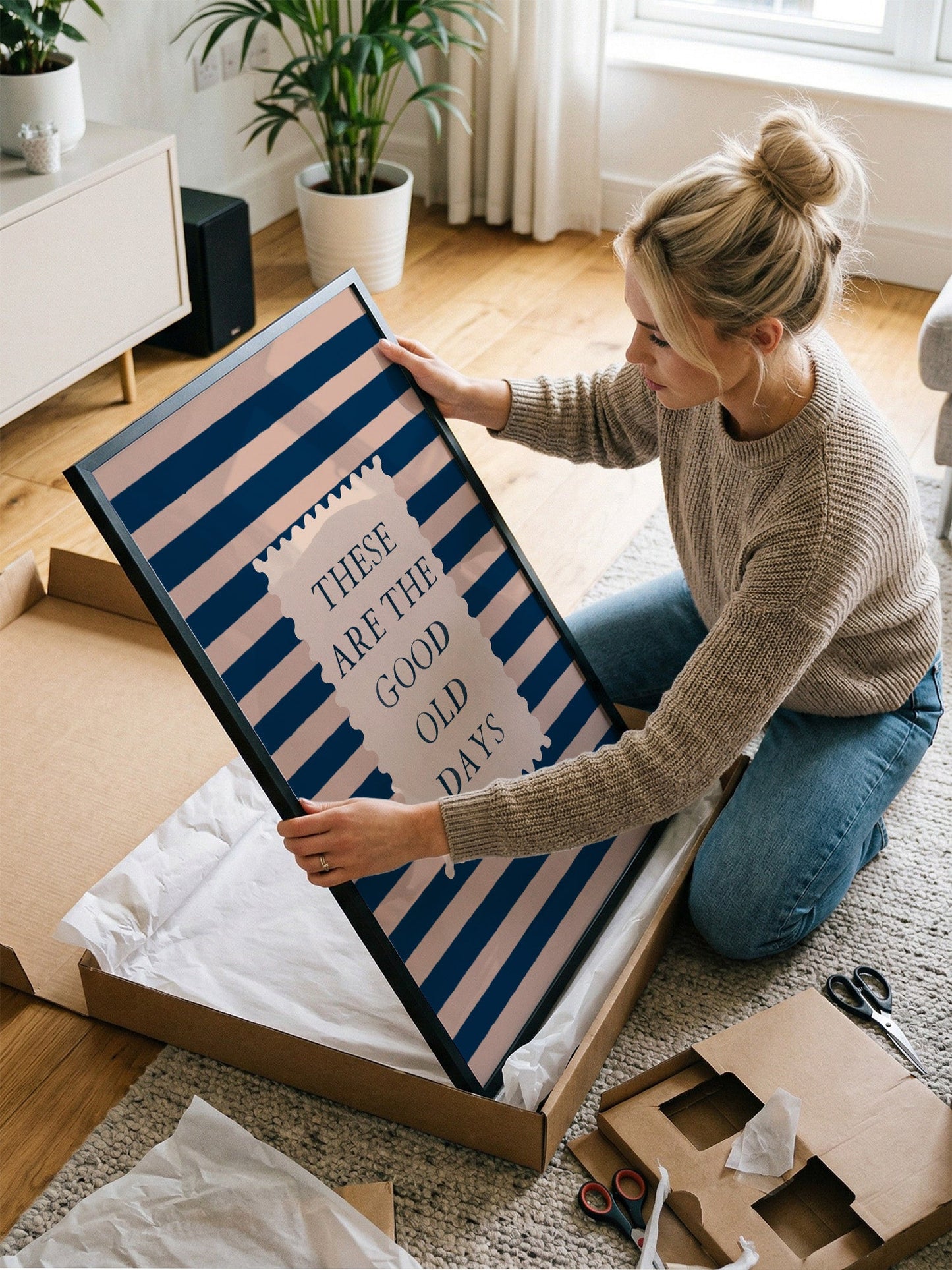 A woman is kneeling on the floor and holding a framed poster with the text "These are the good old days" on it.