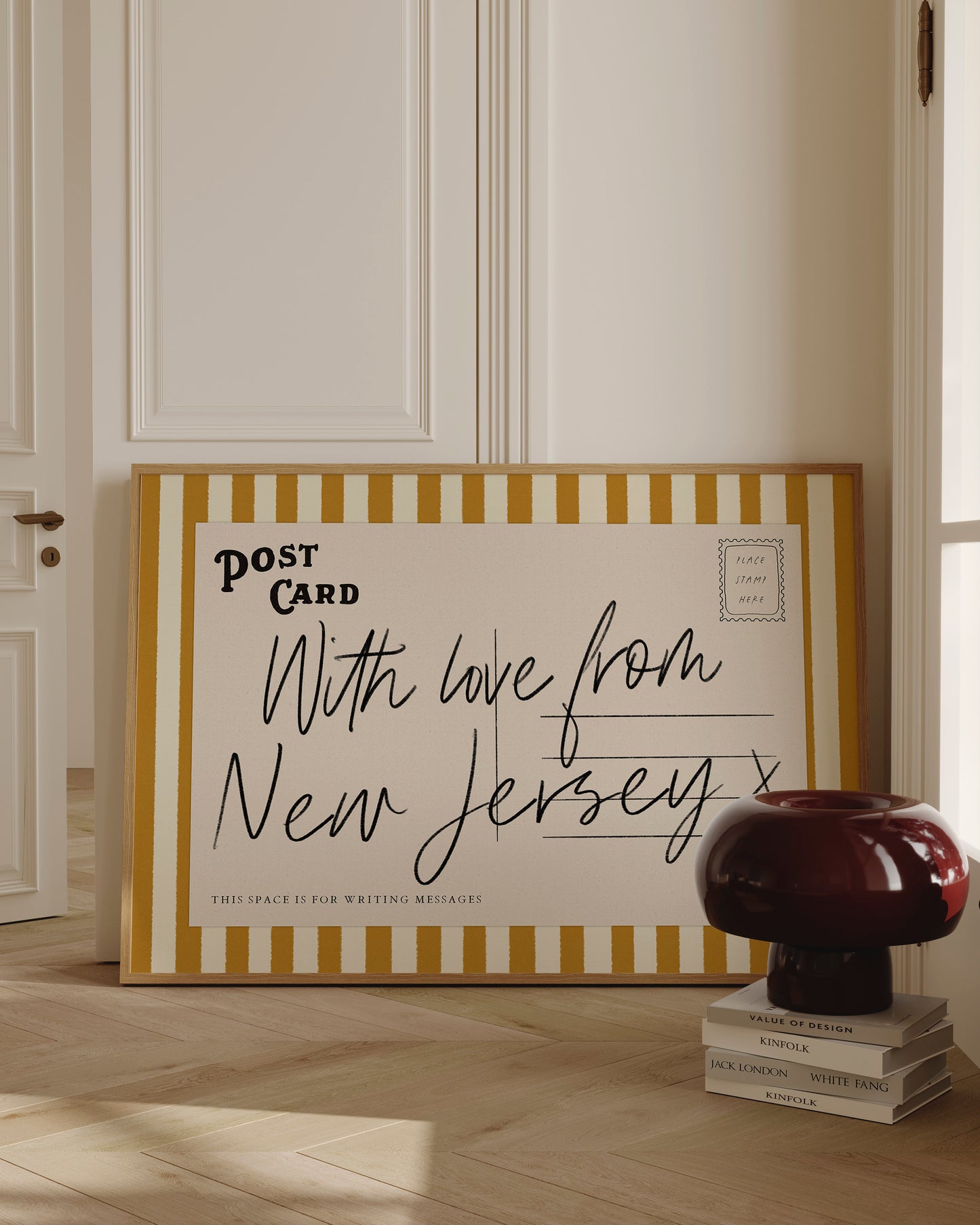 A large framed postcard with handwritten text, placed on a wooden floor next to a stack of books and a lamp.
