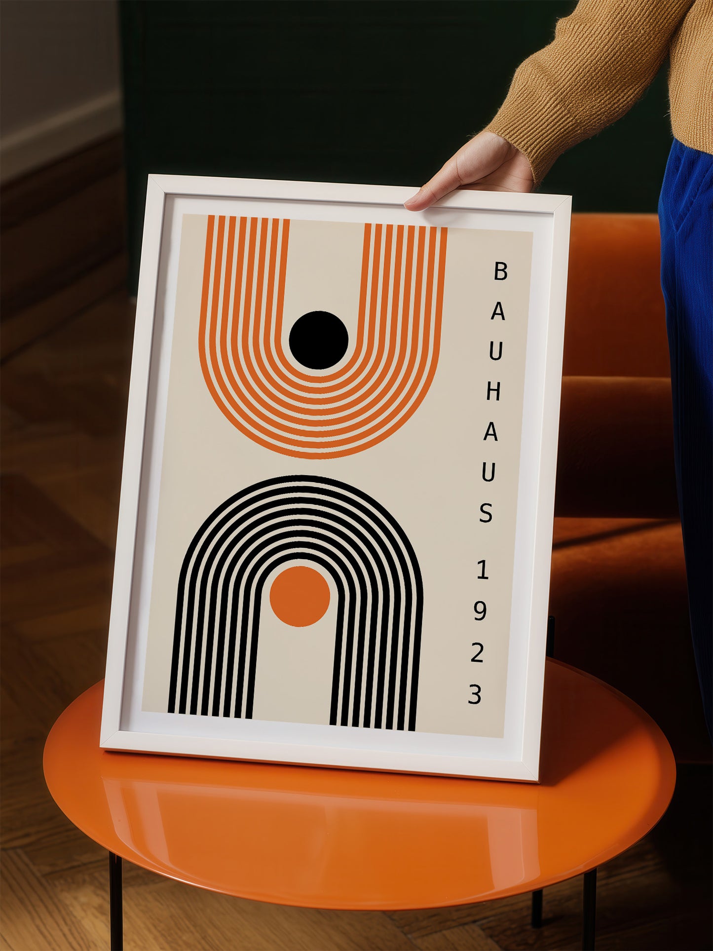 A person is holding a framed poster with a minimalist design featuring abstract shapes and lines in orange, black, and white.