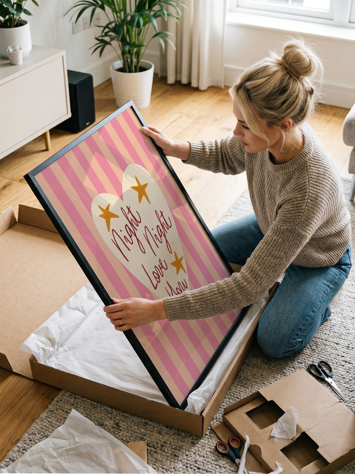 A woman is kneeling on the floor and holding a large framed picture with a pink and white striped background and a heart design.