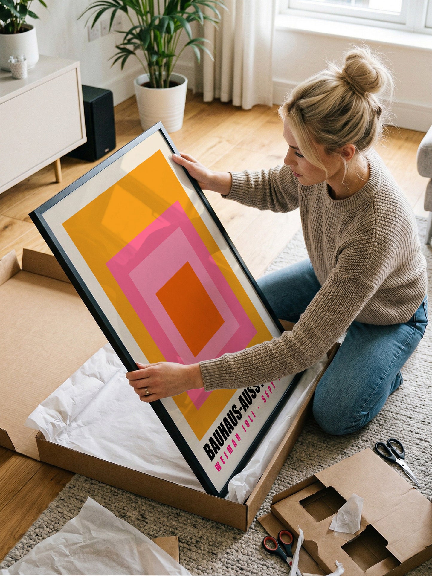 A woman is kneeling on the floor and carefully handling a large framed print, which appears to be a piece of artwork.