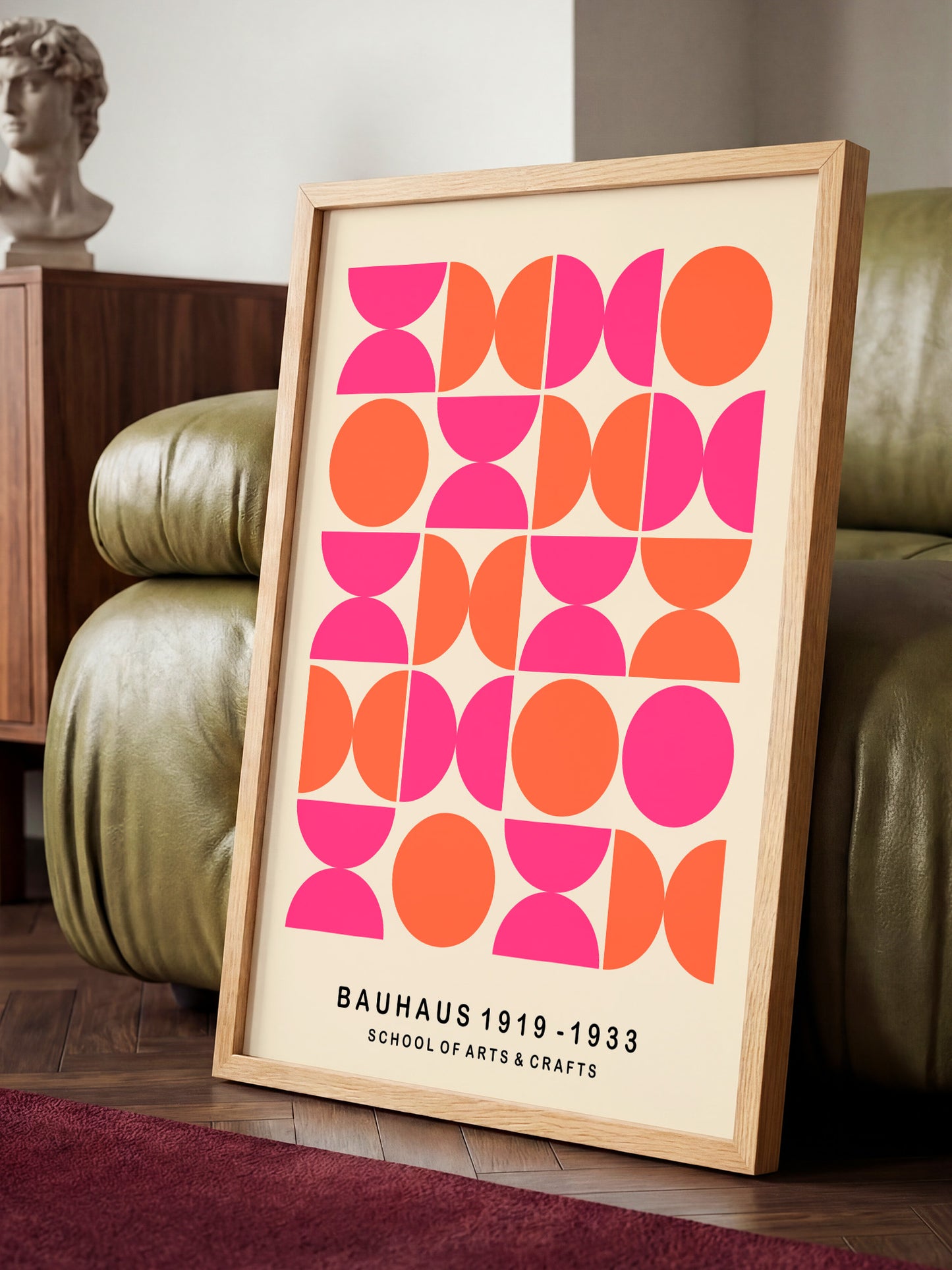 A framed poster with a geometric design in pink and orange shapes, sitting on a wooden floor in front of a green leather couch.