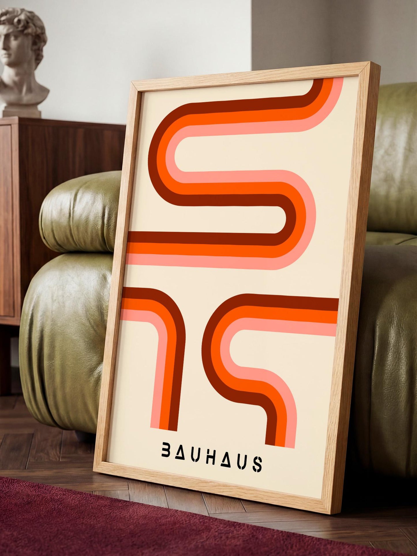 a framed poster with a bold, abstract design in shades of orange and pink, sitting on a wooden floor in front of a green leather couch.