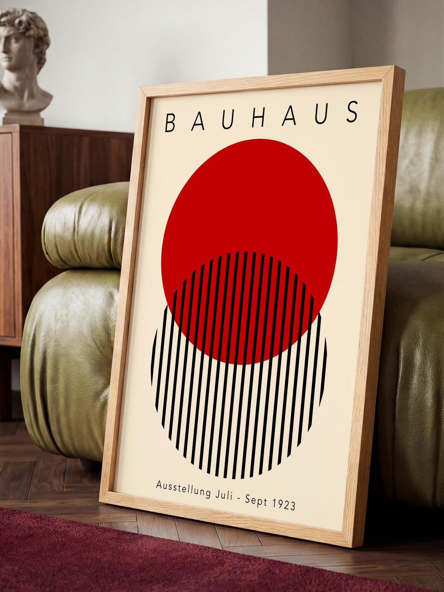 a framed poster of the Bauhaus art school, with a bold red circle and black stripes as the central design element. The poster is displayed on a wooden floor, with a green leather couch visible in the background.