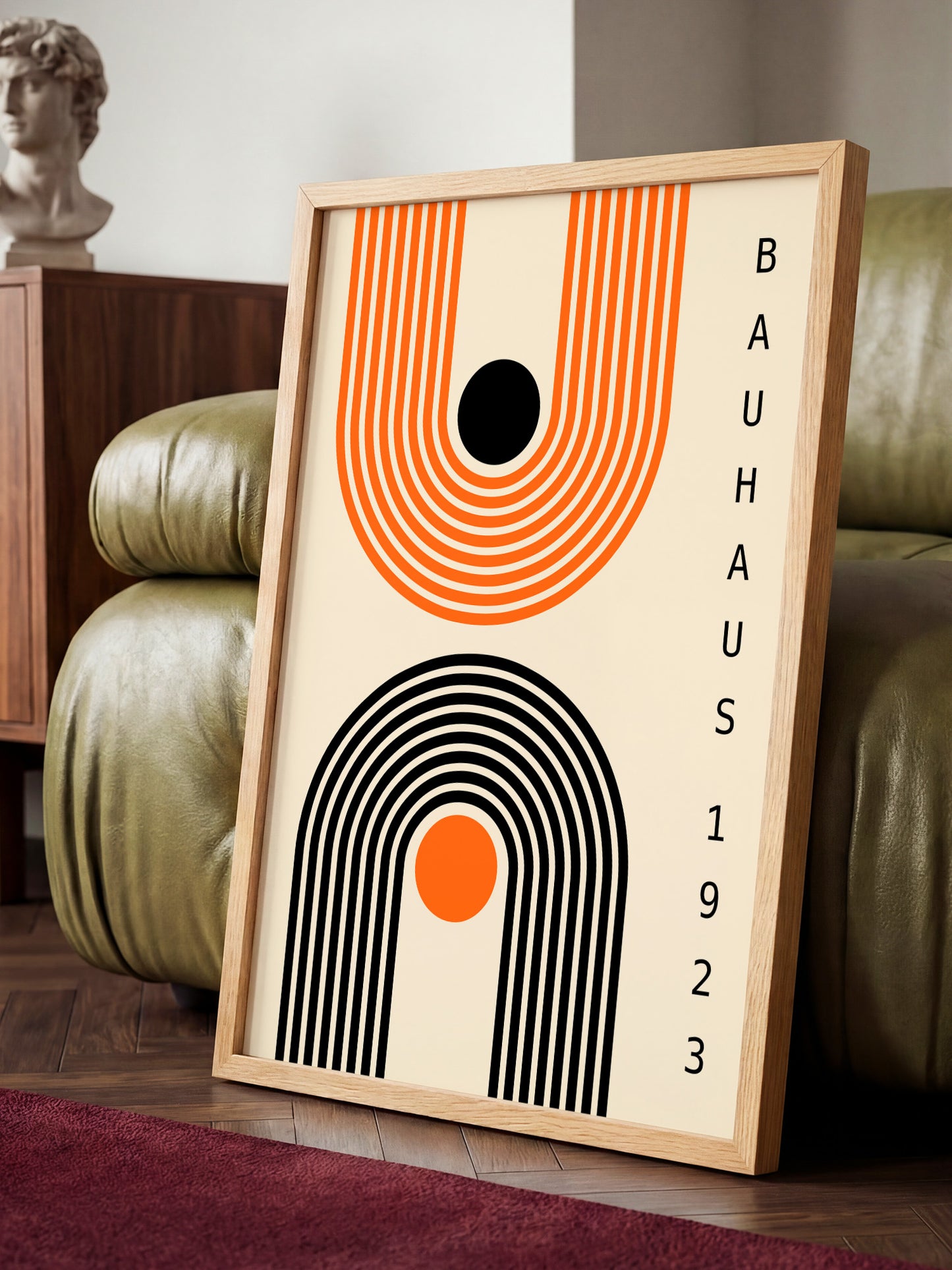 a framed poster with abstract geometric designs in black, orange, and white colors, placed on a wooden floor in front of a green leather couch.