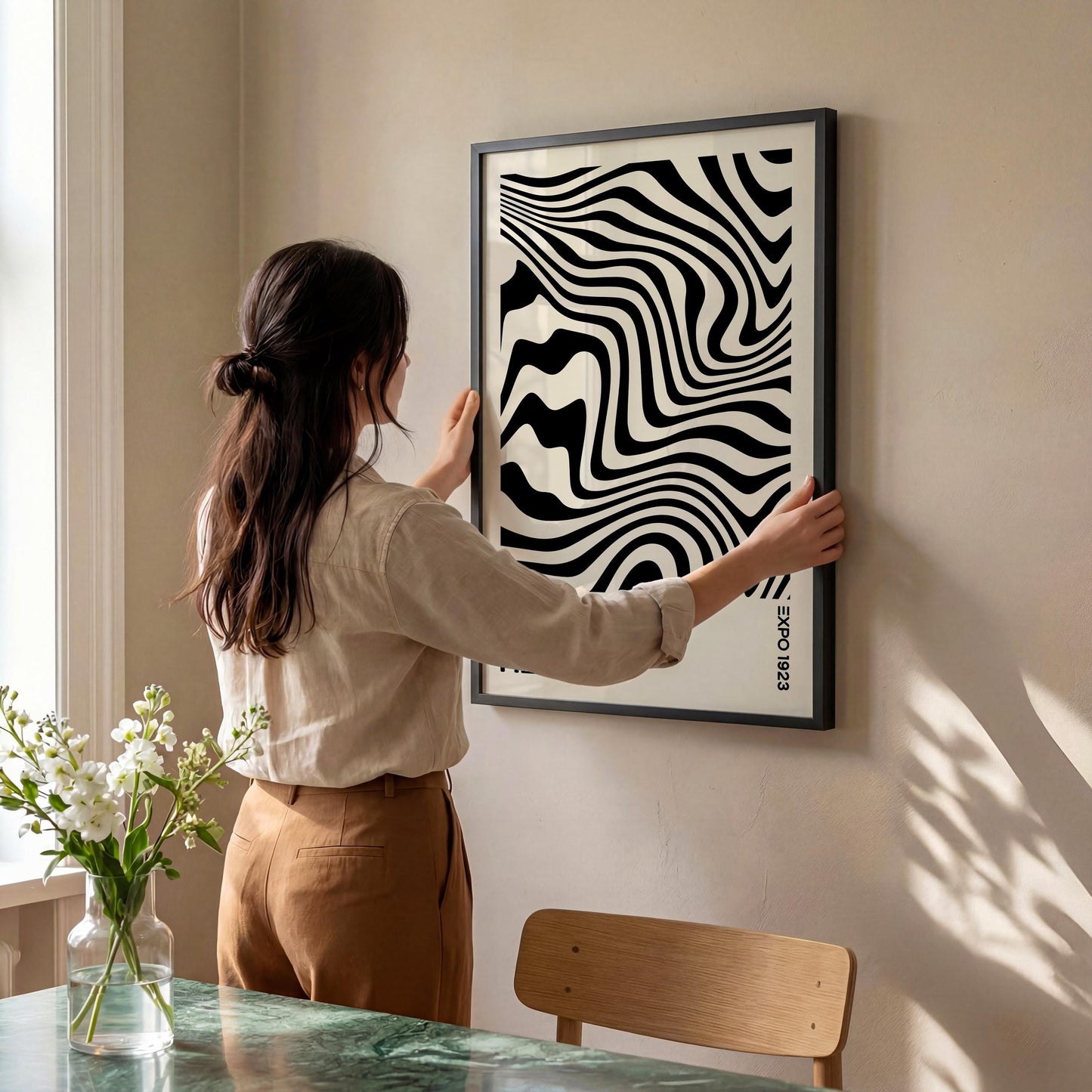 A woman is hanging a framed poster with a black and white abstract design on a wall in a room with a dining table and chairs.