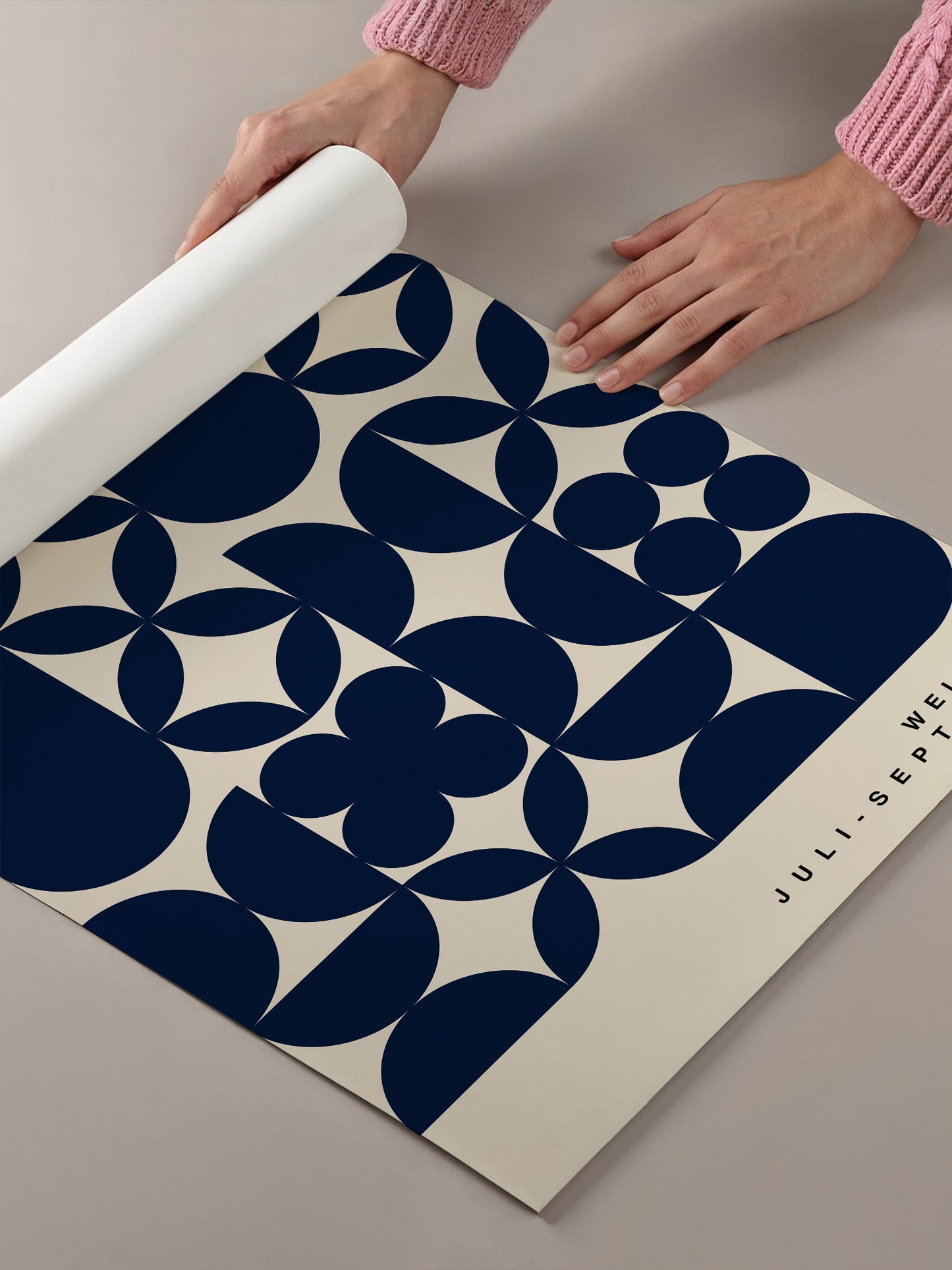 A person's hands are holding a roll of paper, which appears to be a patterned paper, and they are placing it on a table.