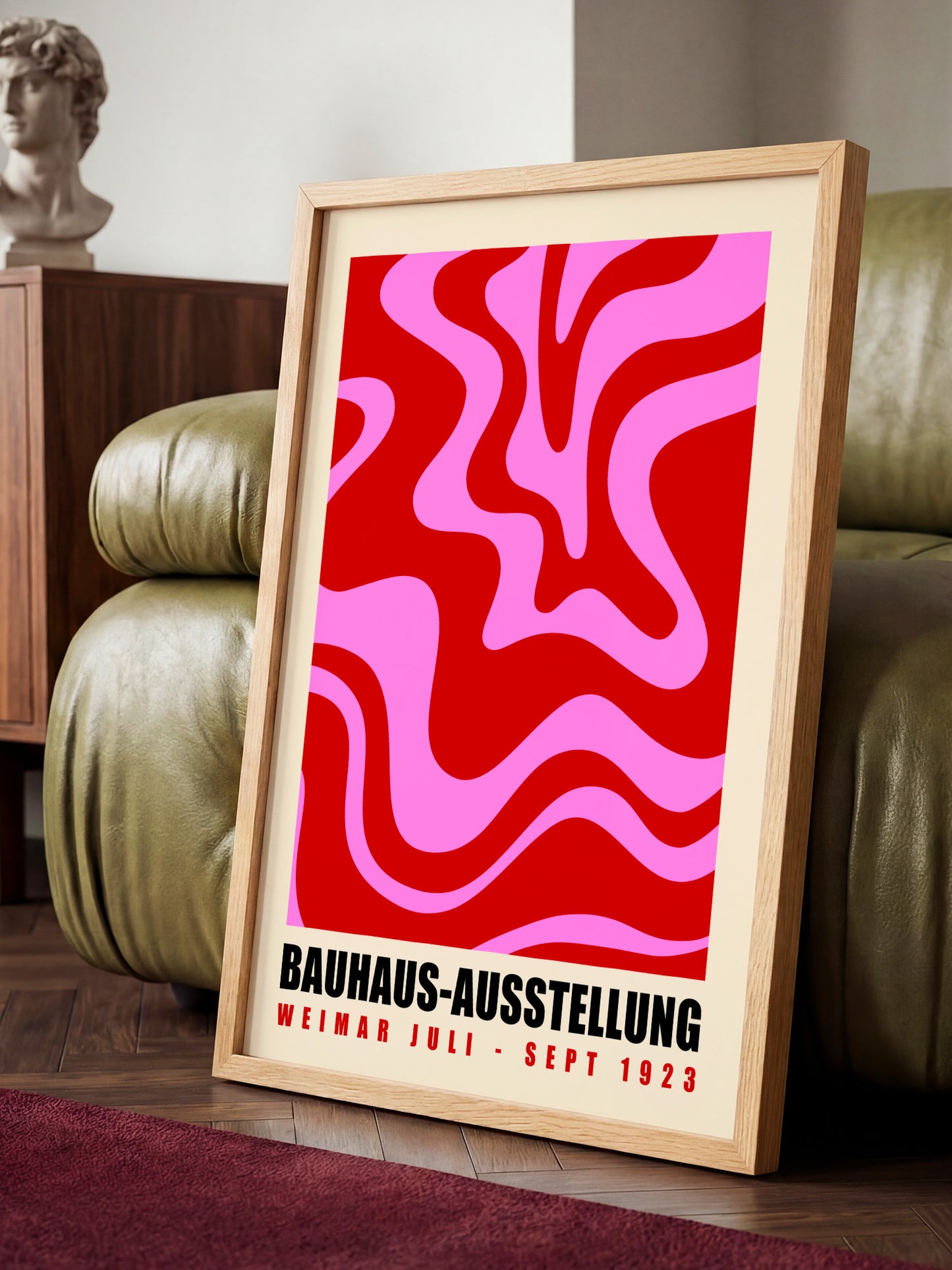 A framed poster with a bold, abstract design in red and pink colors, sitting on a wooden floor in a room with a green leather couch.