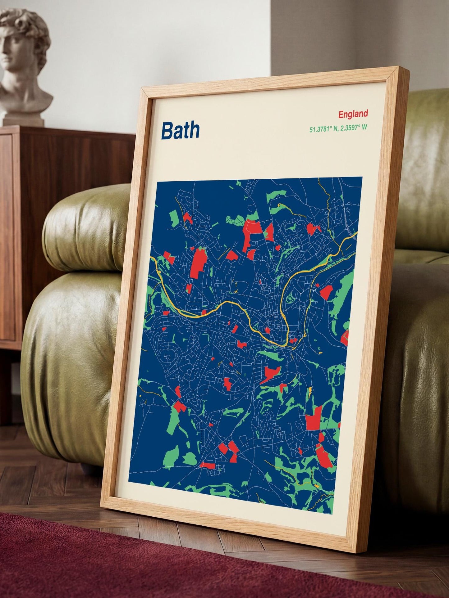 A framed map of Bath, England, with a blue background and red and green accents, is displayed on a wooden floor in a living room setting.
