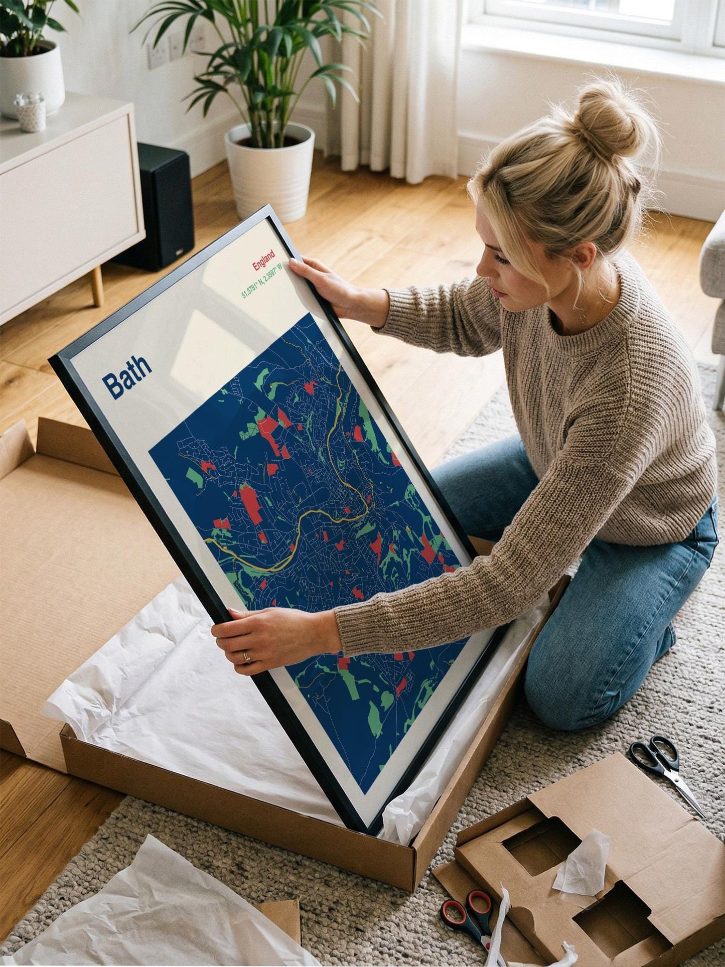 A woman is kneeling on the floor and holding a framed map of Bath, England, which is placed in a cardboard box.