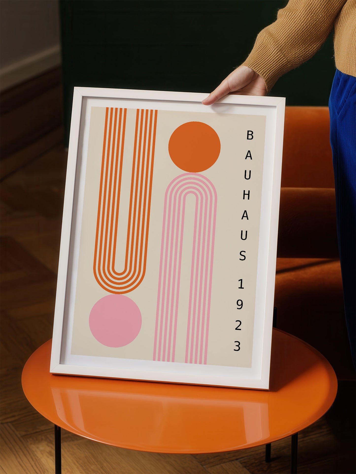 A person is holding a framed poster with a minimalist design featuring geometric shapes in shades of orange and pink.