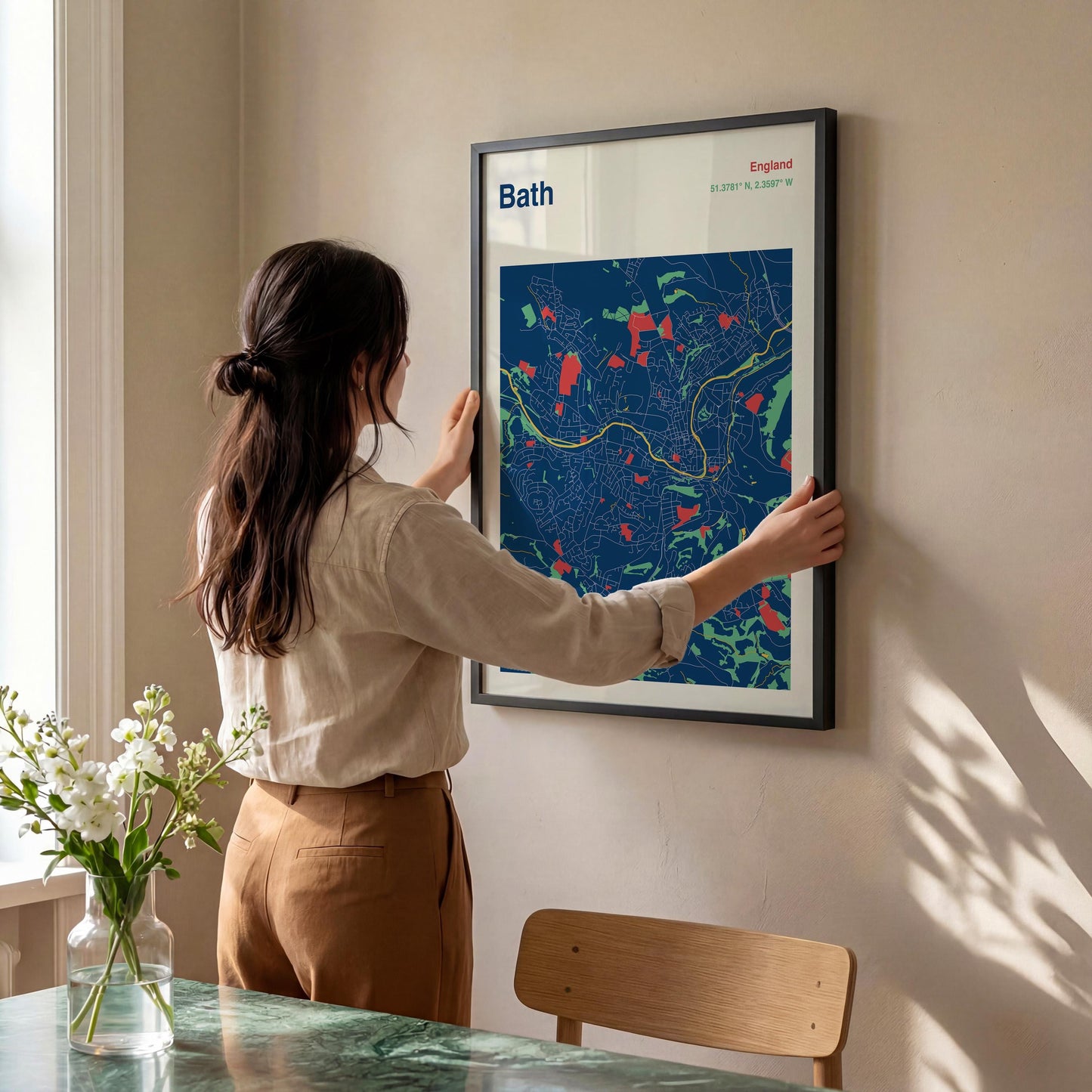 A woman is hanging a framed poster of a map of Bath, England, on a wall in a room with a dining table and a vase of flowers.