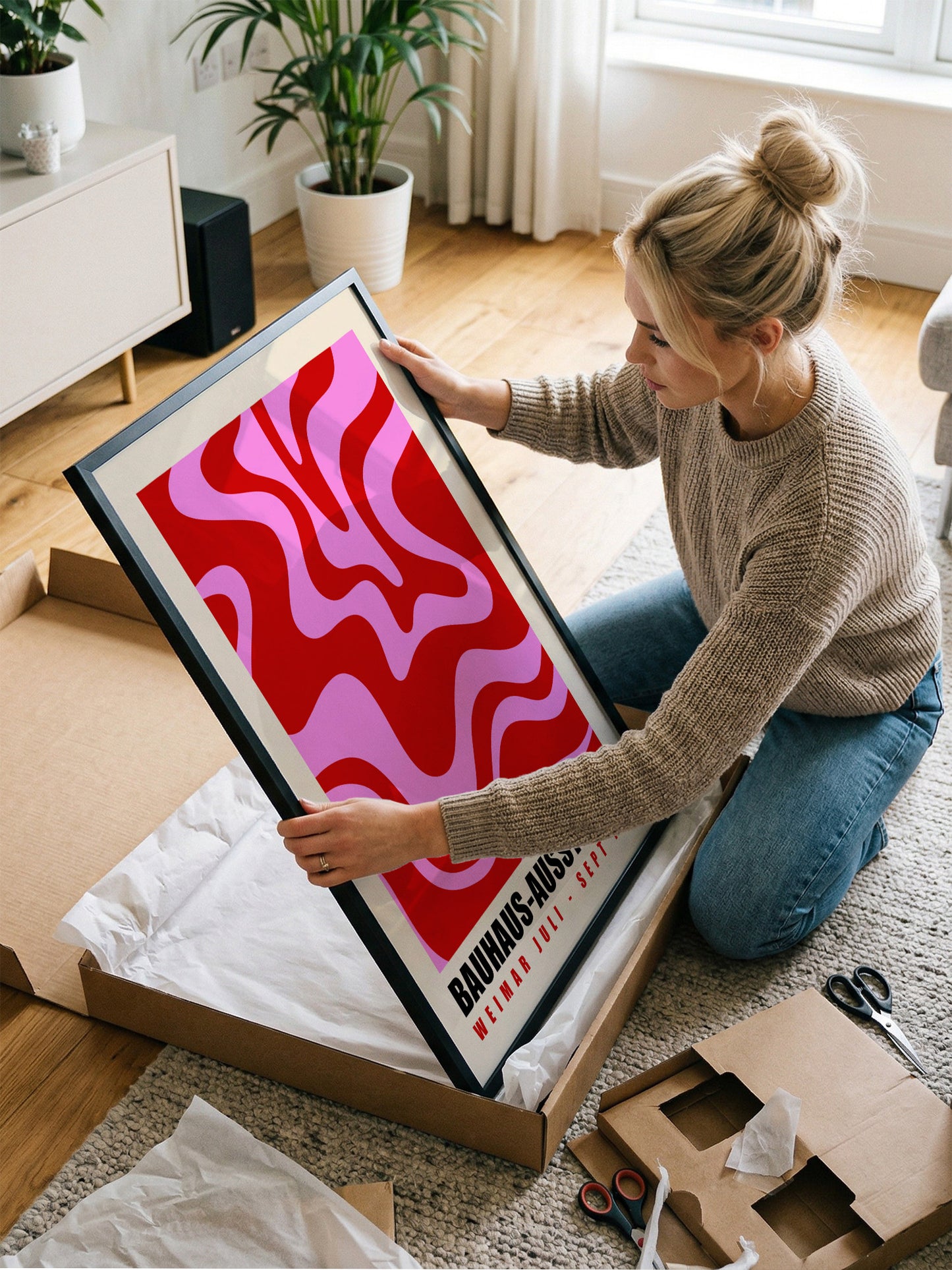 A woman is kneeling on the floor and carefully handling a large framed poster with a striking red and pink abstract design.