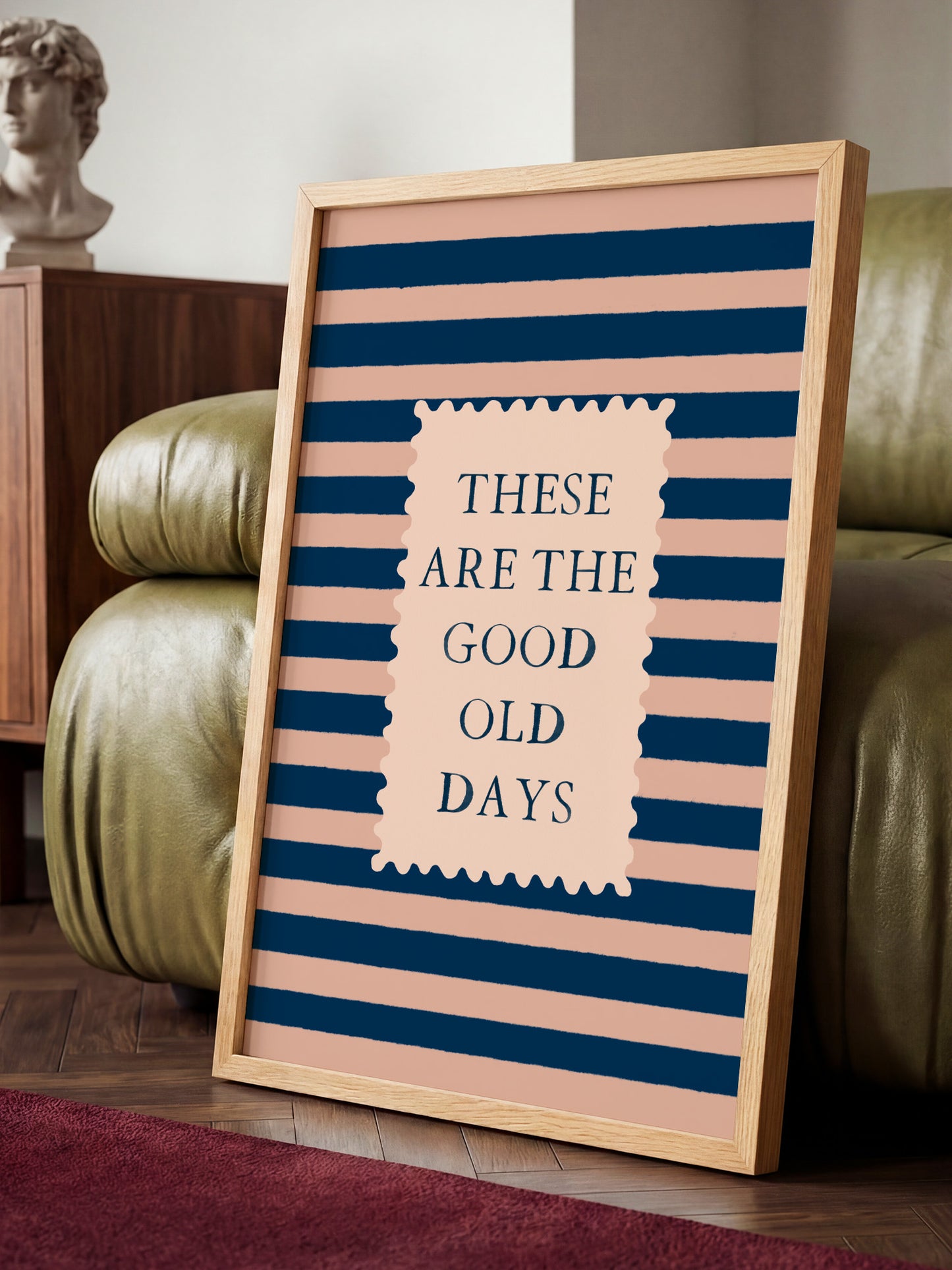 A framed poster with the text "These are the good old days" displayed on it, standing on a wooden floor in front of a green leather couch.