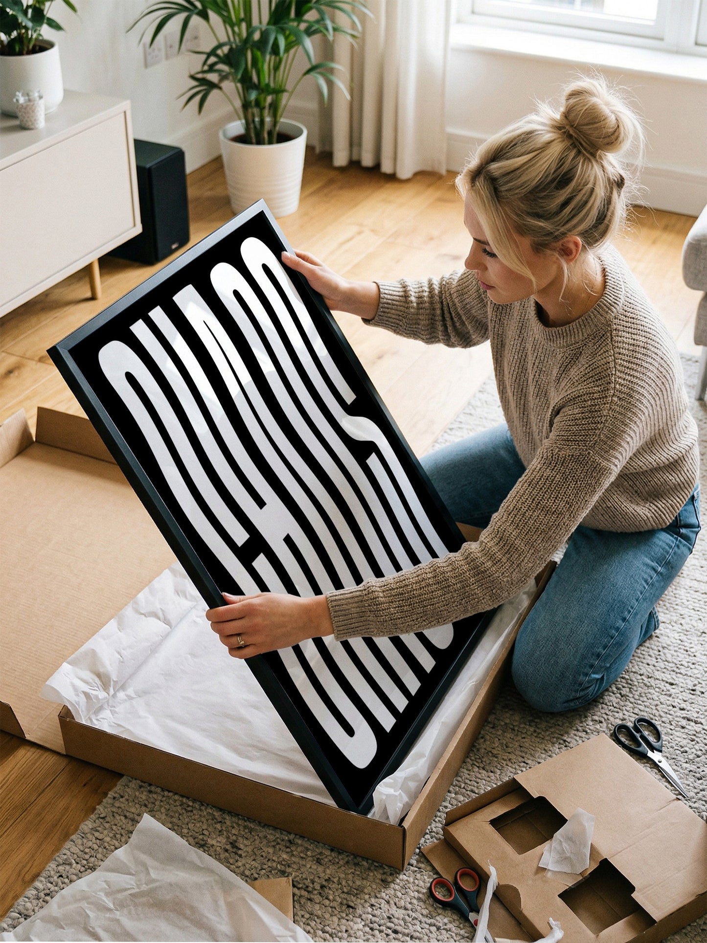 A woman is kneeling on the floor and carefully opening a cardboard box, which contains a large black and white framed print.