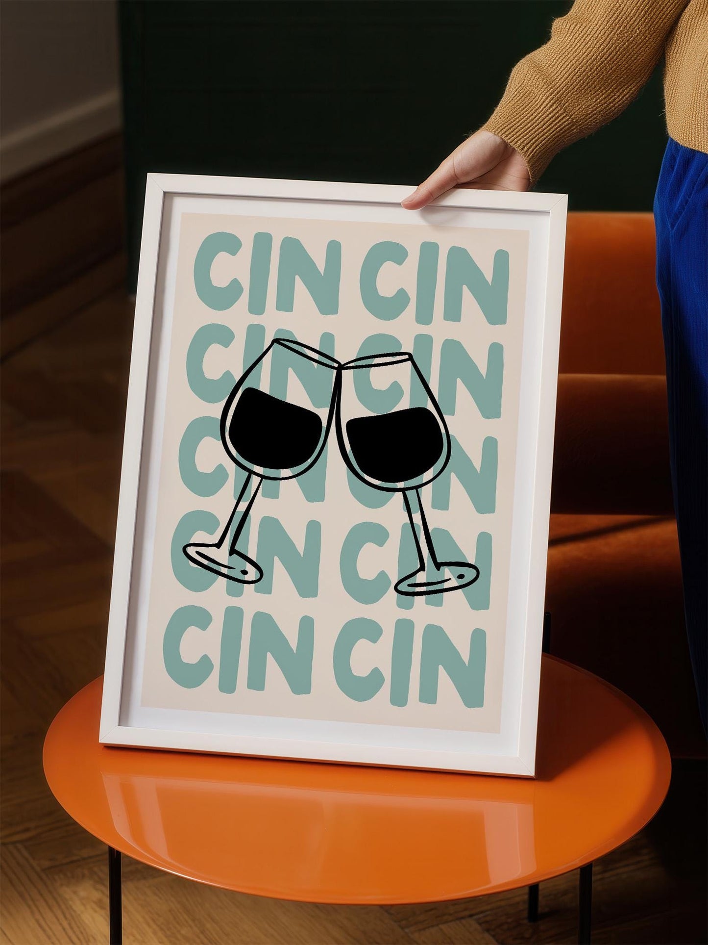 A person is holding up a framed print with a graphic design featuring two wine glasses with the word "CIN" repeated in a stylized font.