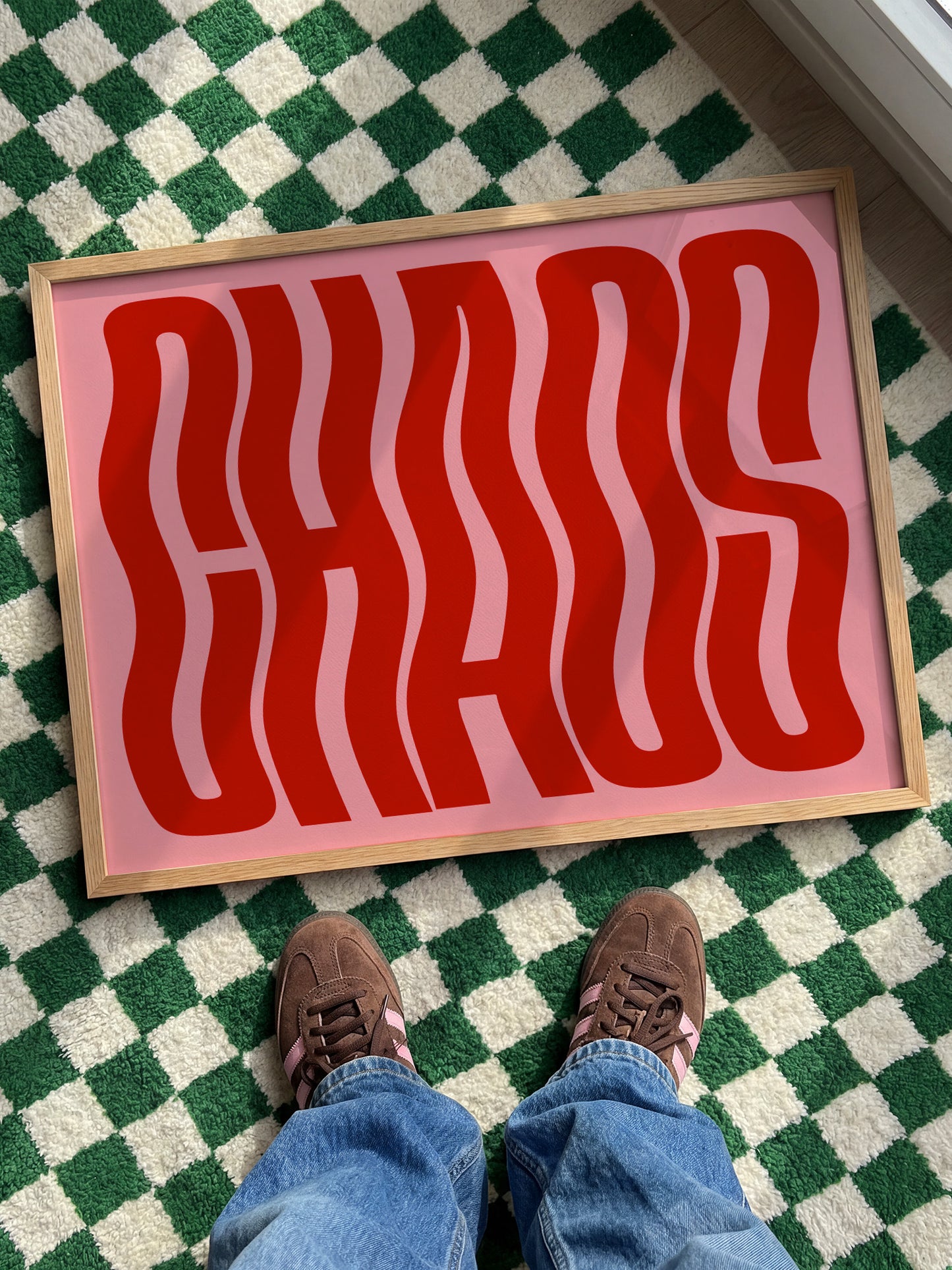 A pair of feet standing on a checkered green and white rug, with a large red text poster or sign with the word "CHAOS" displayed on it.