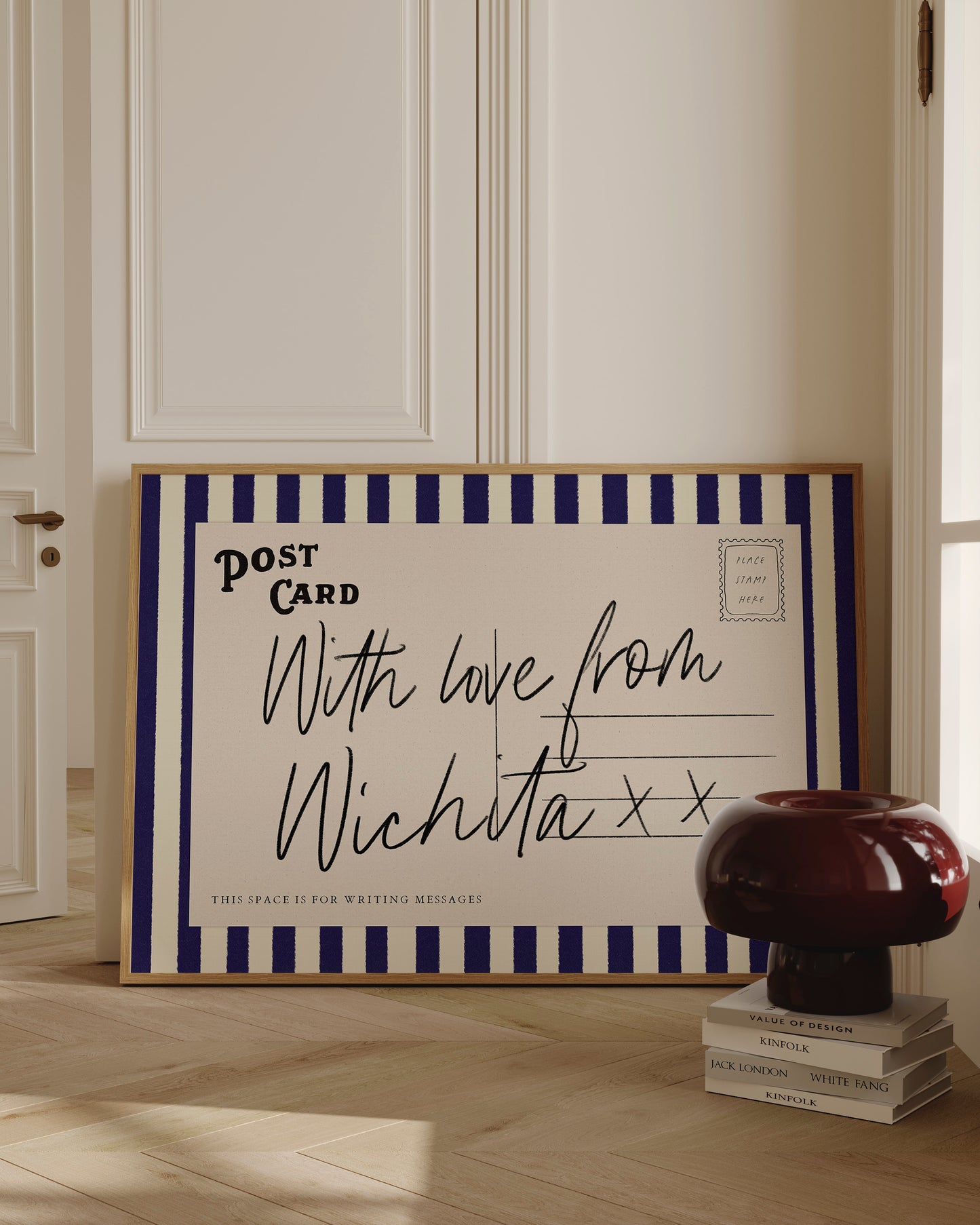 A framed postcard with a handwritten message is displayed on a wooden floor, next to a stack of books and a lamp.