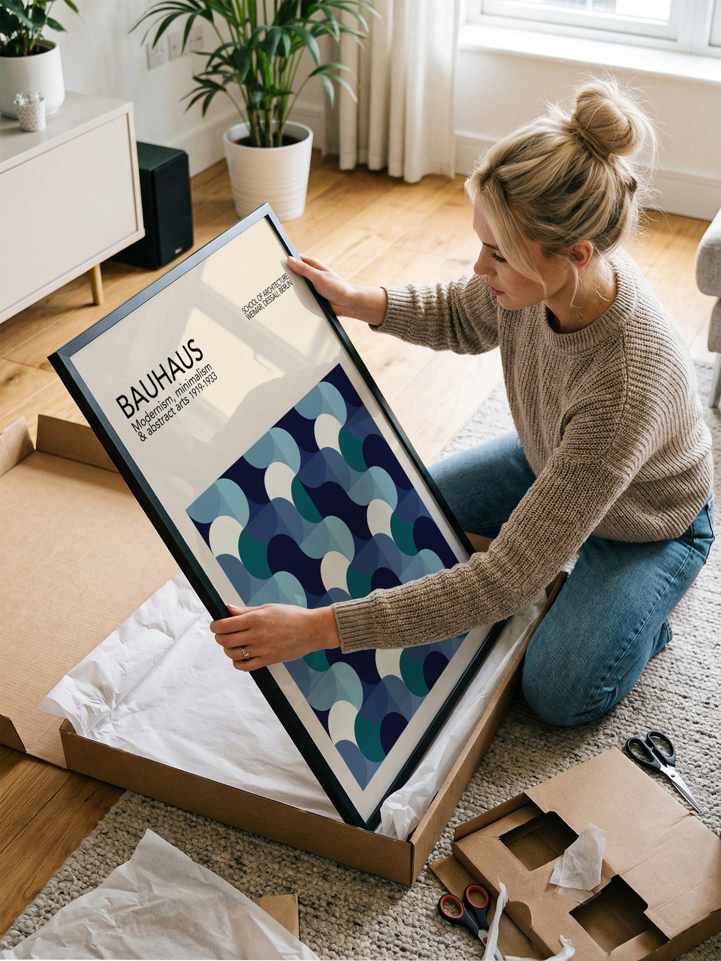 A woman is kneeling on the floor and carefully opening a large cardboard box, which contains a framed poster of the Bauhaus art style.