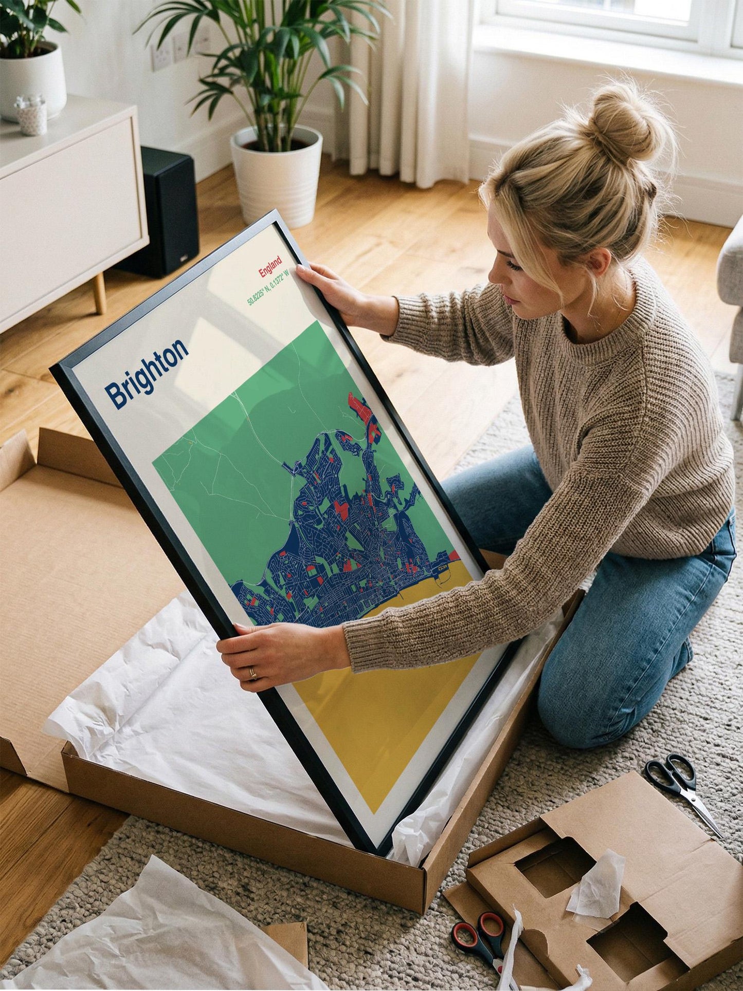 A woman is kneeling on the floor and holding a framed map of Brighton, which is a city in the United Kingdom.