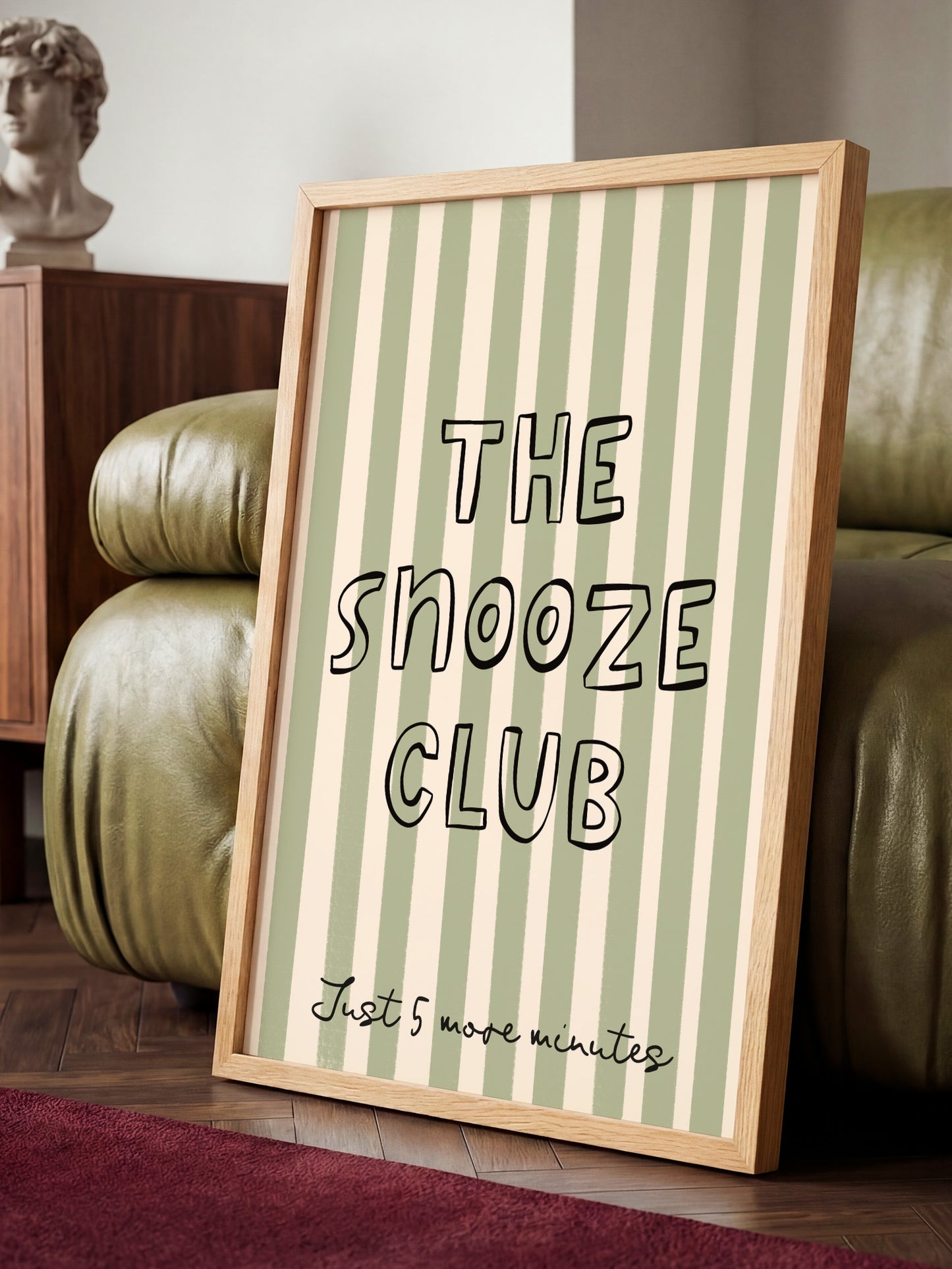 A framed poster with the text "THE SNOOZE CLUB" and "Just 5 more minutes" is displayed on a wooden floor in front of a green leather couch.