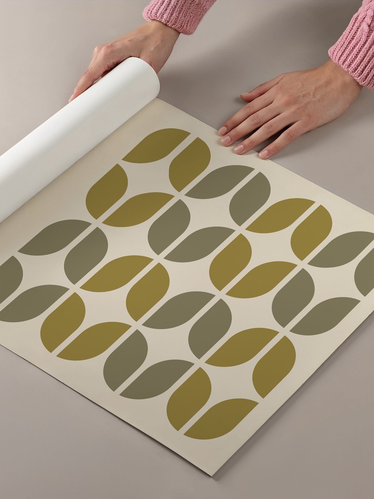 A person's hands are rolling out a roll of wallpaper with a repeating pattern of green and brown circles on a light-colored background.