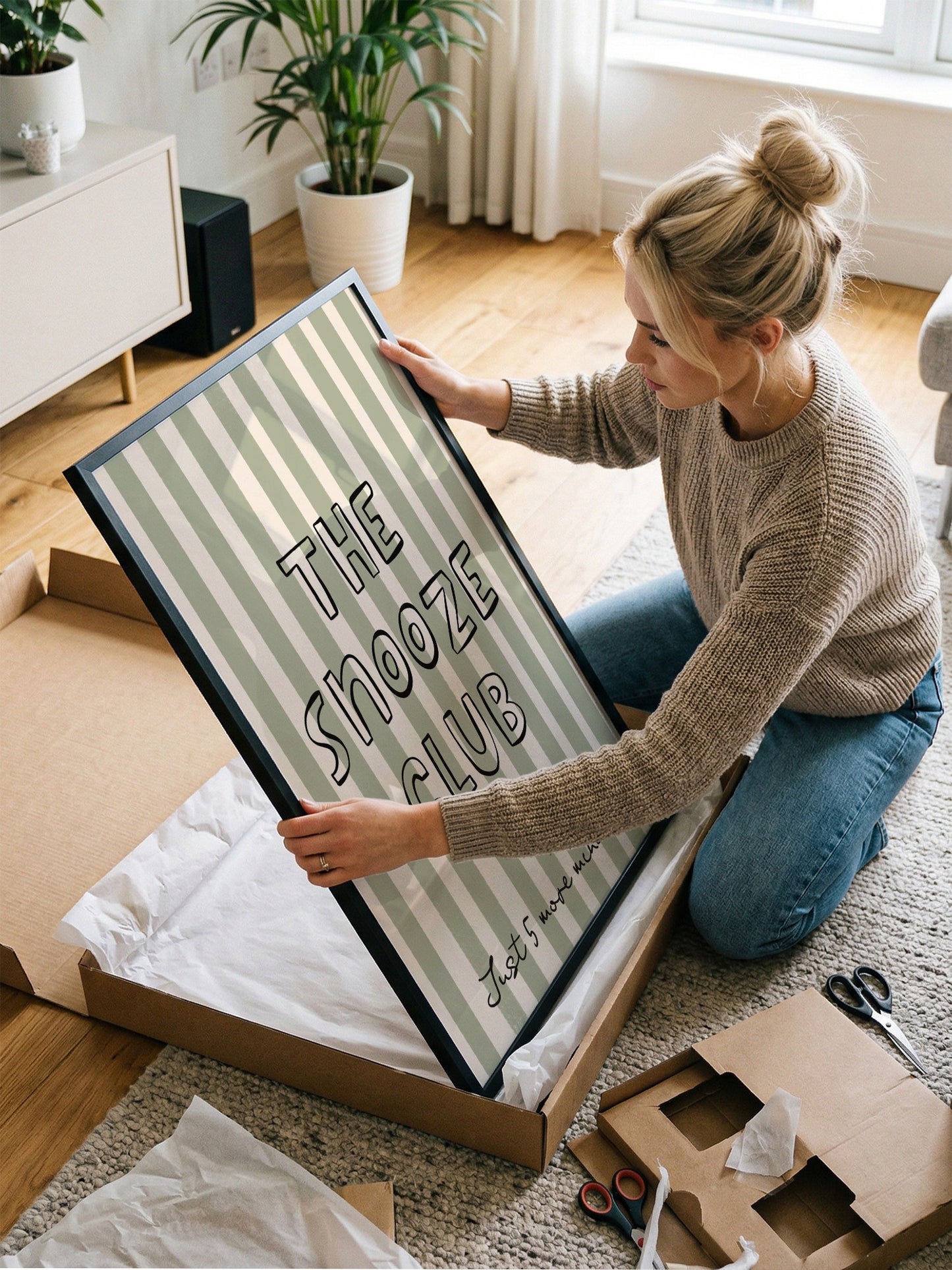 A woman is kneeling on the floor and holding a framed poster with the text "The Shooze Club" on it.