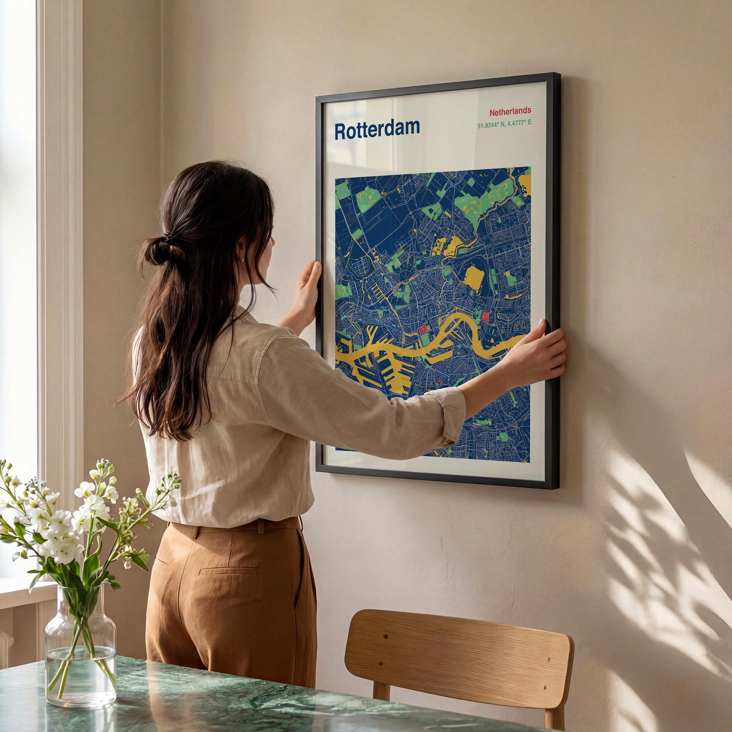 A woman is hanging a framed map of Rotterdam on a wall in a room with a dining table and a vase of flowers.