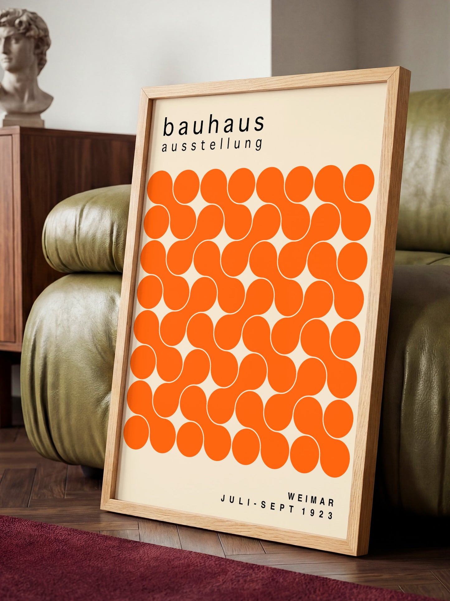 a framed poster with a repeating pattern of orange circles on a beige background, placed on a wooden floor in front of a green leather couch.