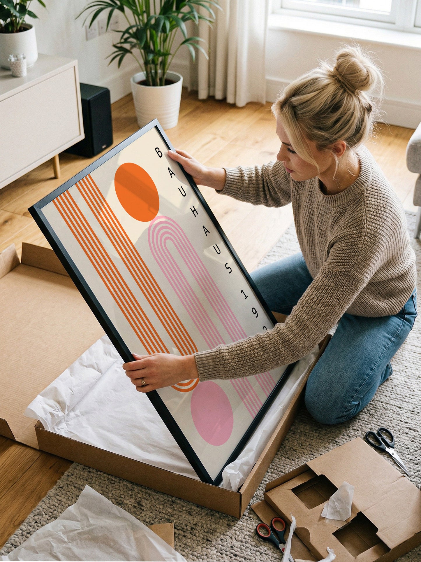 A woman is kneeling on the floor and carefully opening a large cardboard box, which contains a framed piece of art.