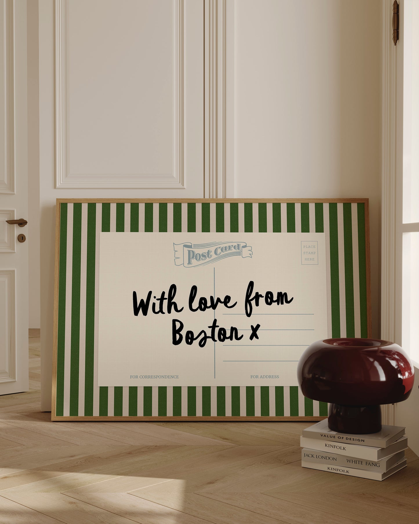 A framed postcard with a green and white striped border, displaying a message that reads "With love from Boston X" and "FOR CORRESPONDENCE". The postcard is placed on a wooden floor, with a stack of books and a lamp nearby.