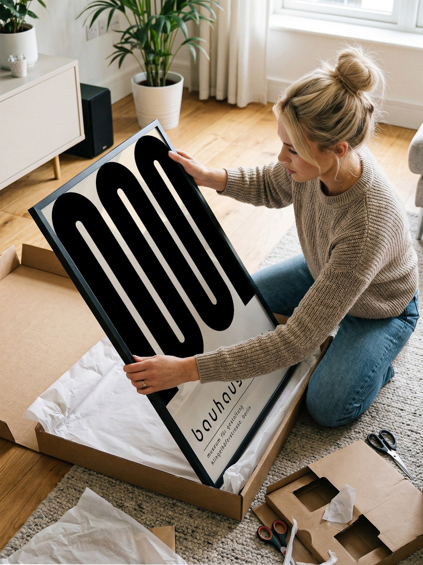 A woman is kneeling on the floor and opening a large cardboard box, which contains a framed poster.