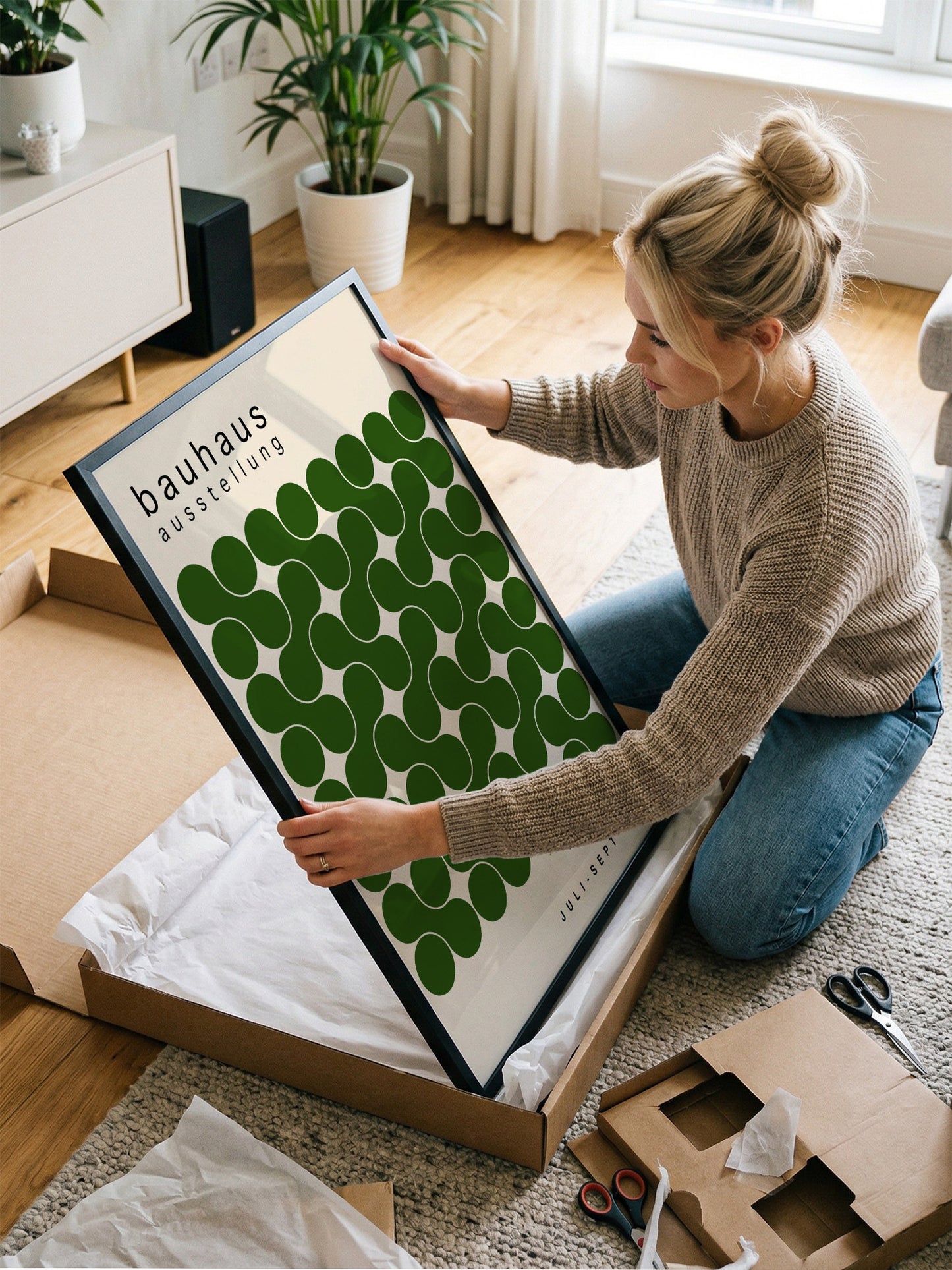 A woman is kneeling on the floor and holding a framed poster with a green and white design.