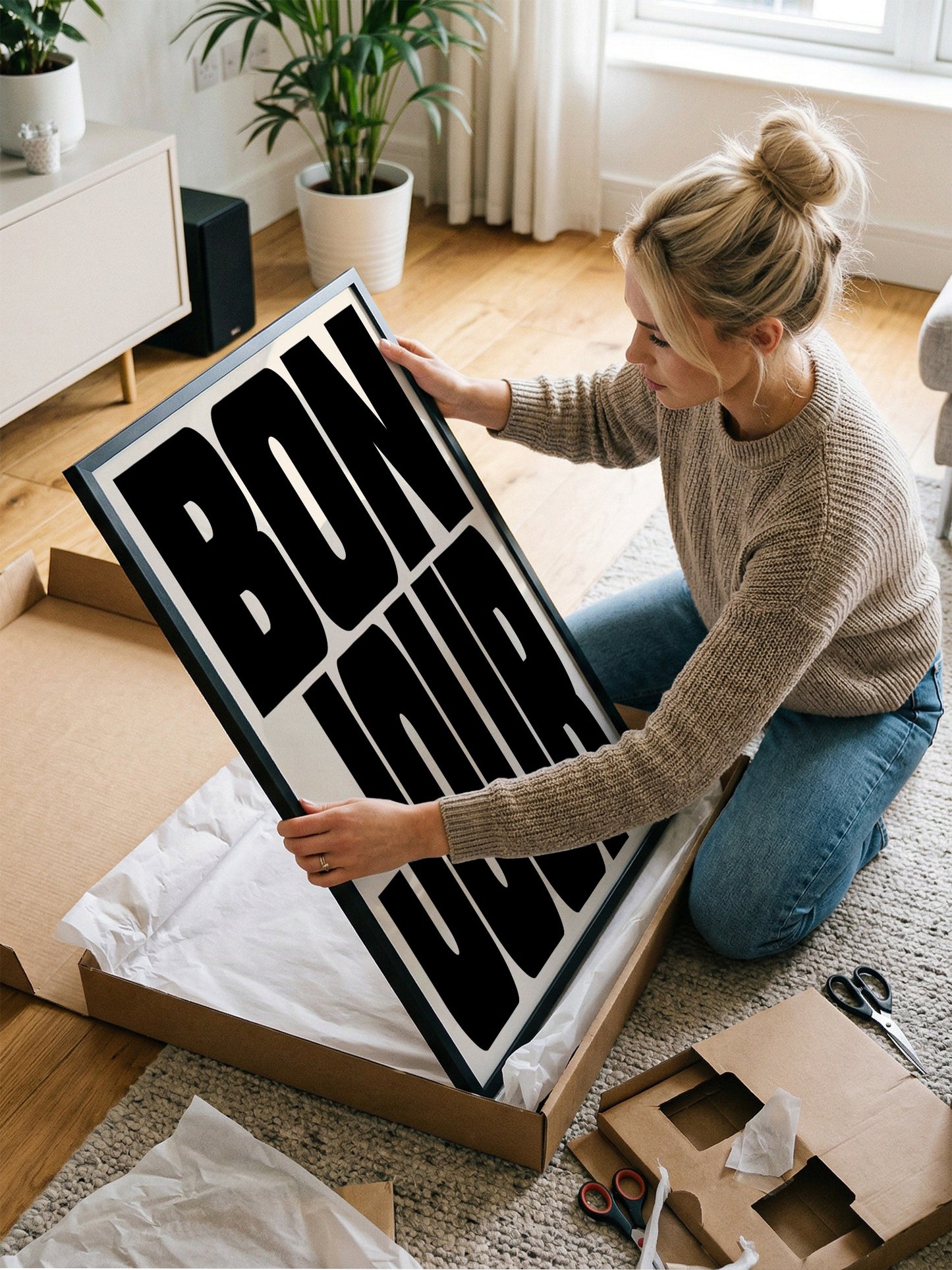 A woman is kneeling on the floor and holding a large framed poster with the word "BON JOUR" written on it.