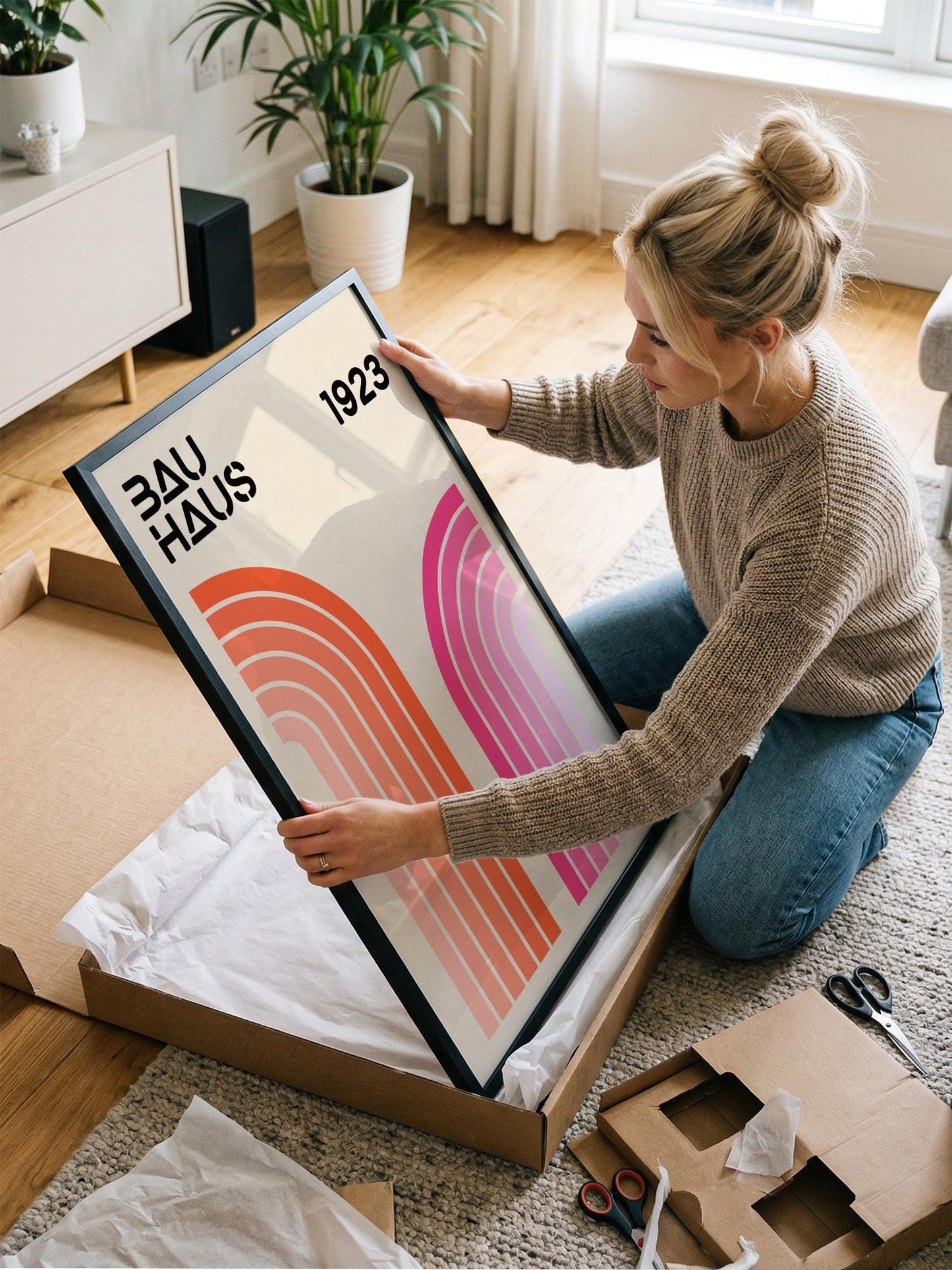 A woman is kneeling on the floor and holding a framed poster with the text "Bauhaus 1923" on it.