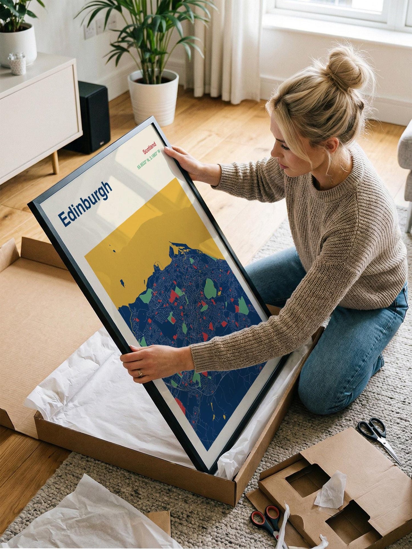 A woman is kneeling on the floor and holding a framed picture of Edinburgh, a city in Scotland.