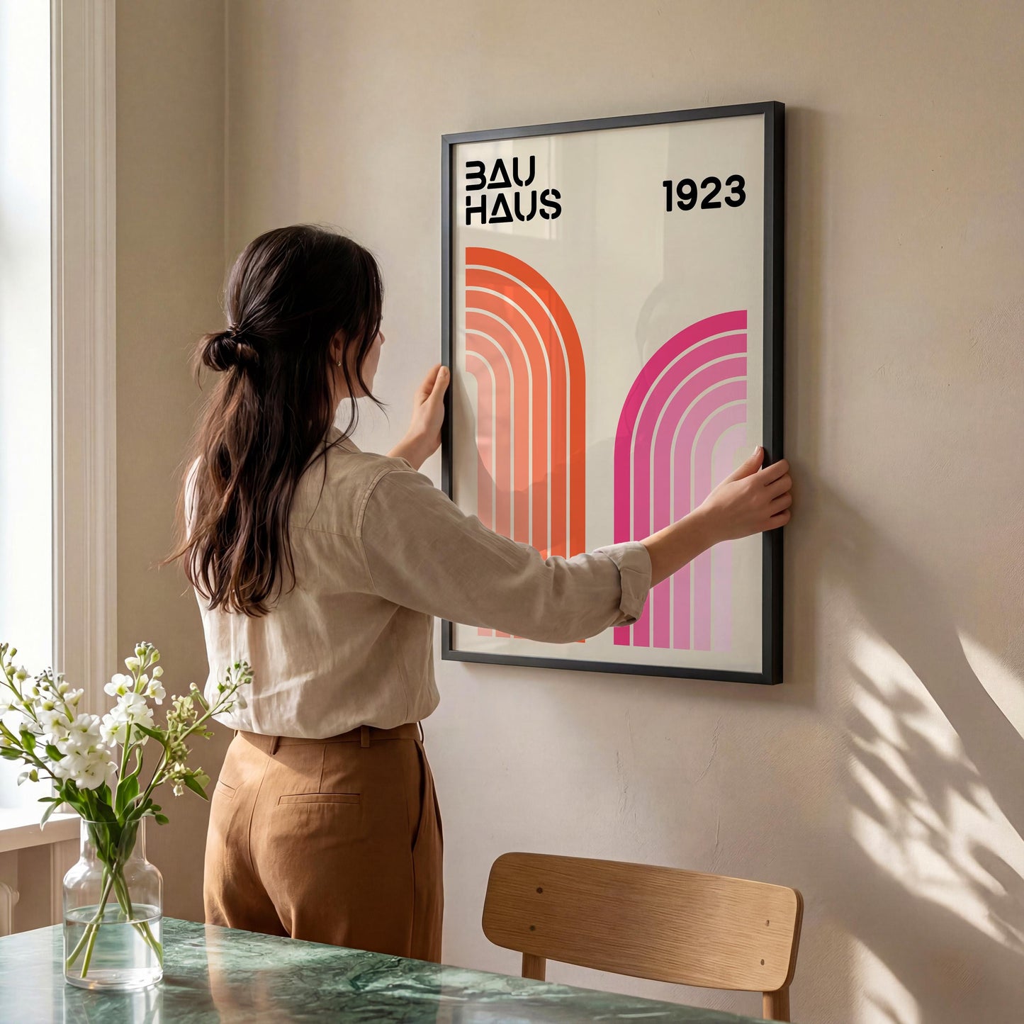 A woman is hanging a framed poster on a wall, which features a design with pink and orange lines.