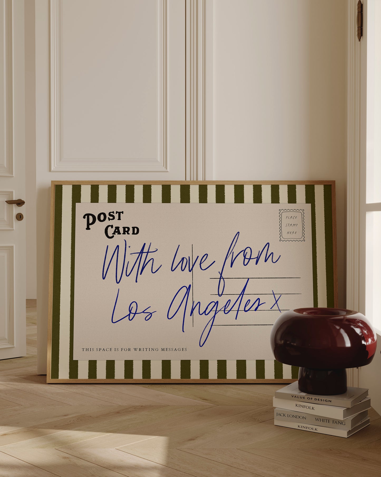 A framed postcard with a handwritten message, placed on a wooden floor next to a stack of books and a lamp.
