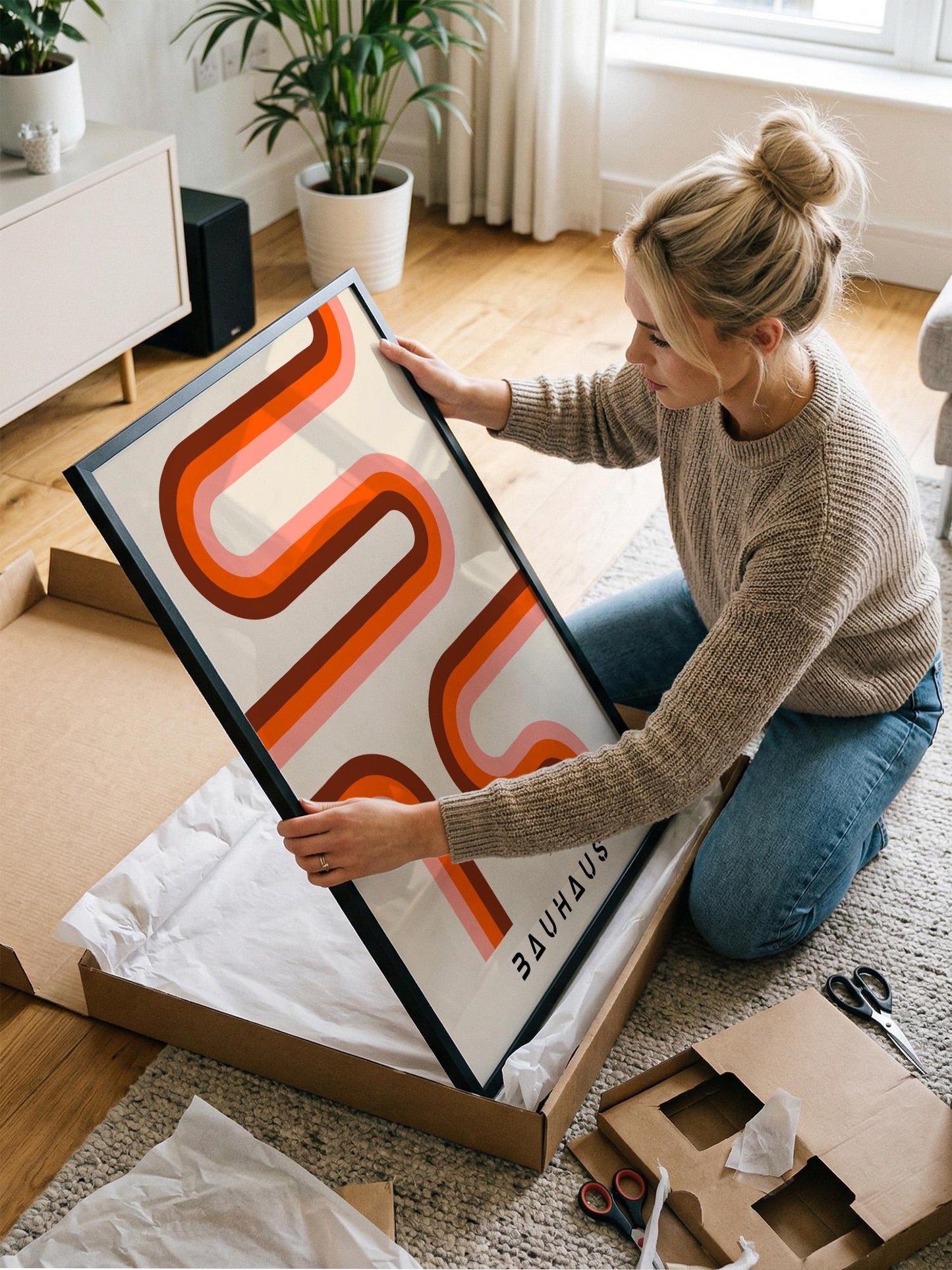 A woman is kneeling on the floor and carefully opening a large picture frame, which has a striking abstract design in orange and red colors.