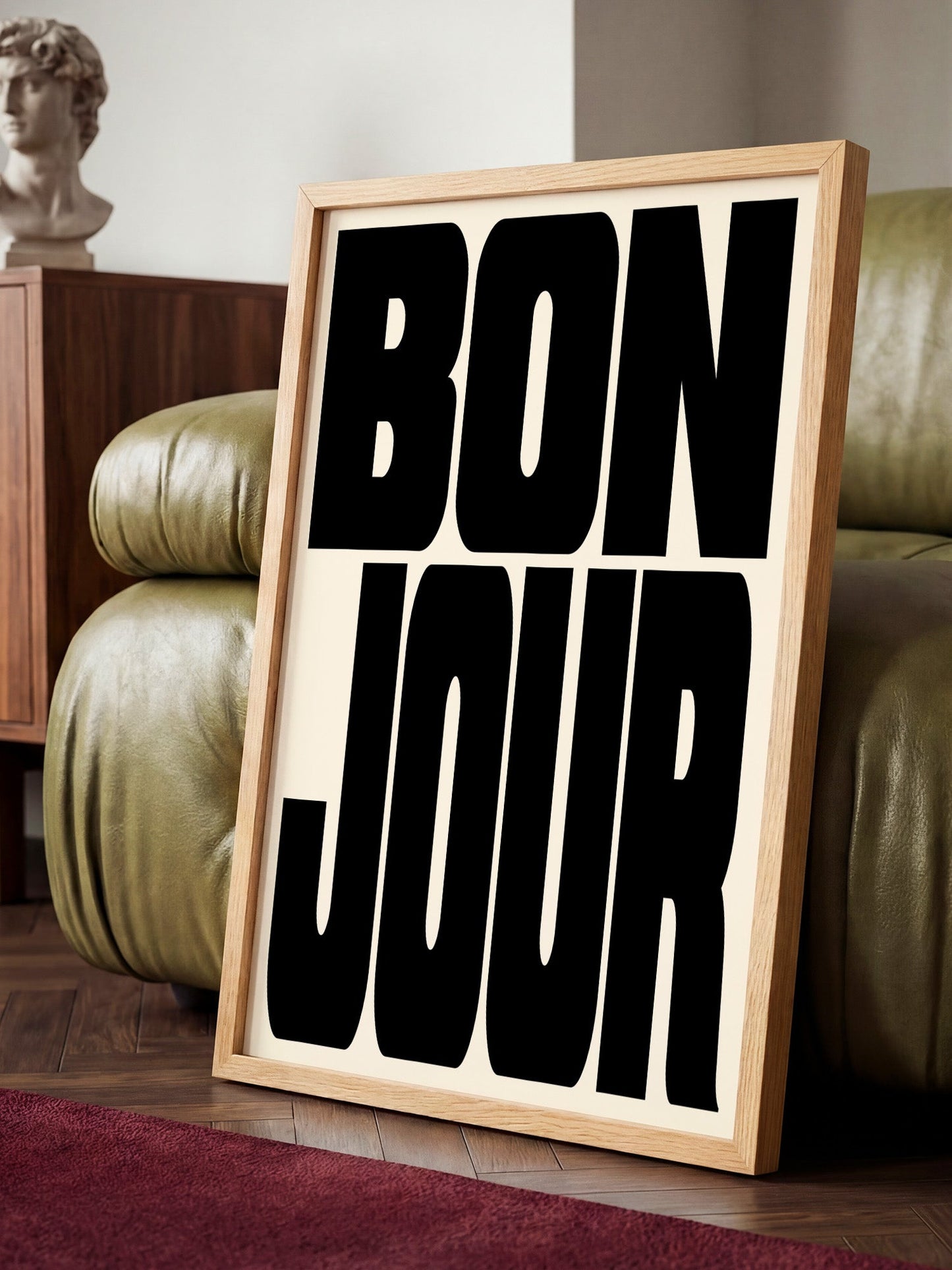 A framed poster with the text " bon jour" in bold black letters is displayed on a wooden floor in a room with a green leather couch.
