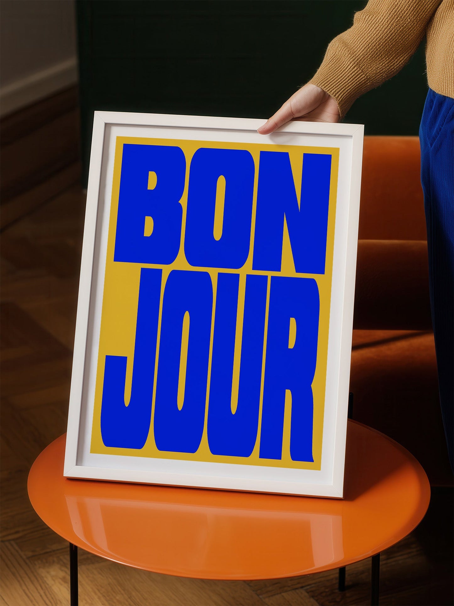 A person is holding up a framed poster with the text "BON JOUR" in large blue letters on a yellow background.