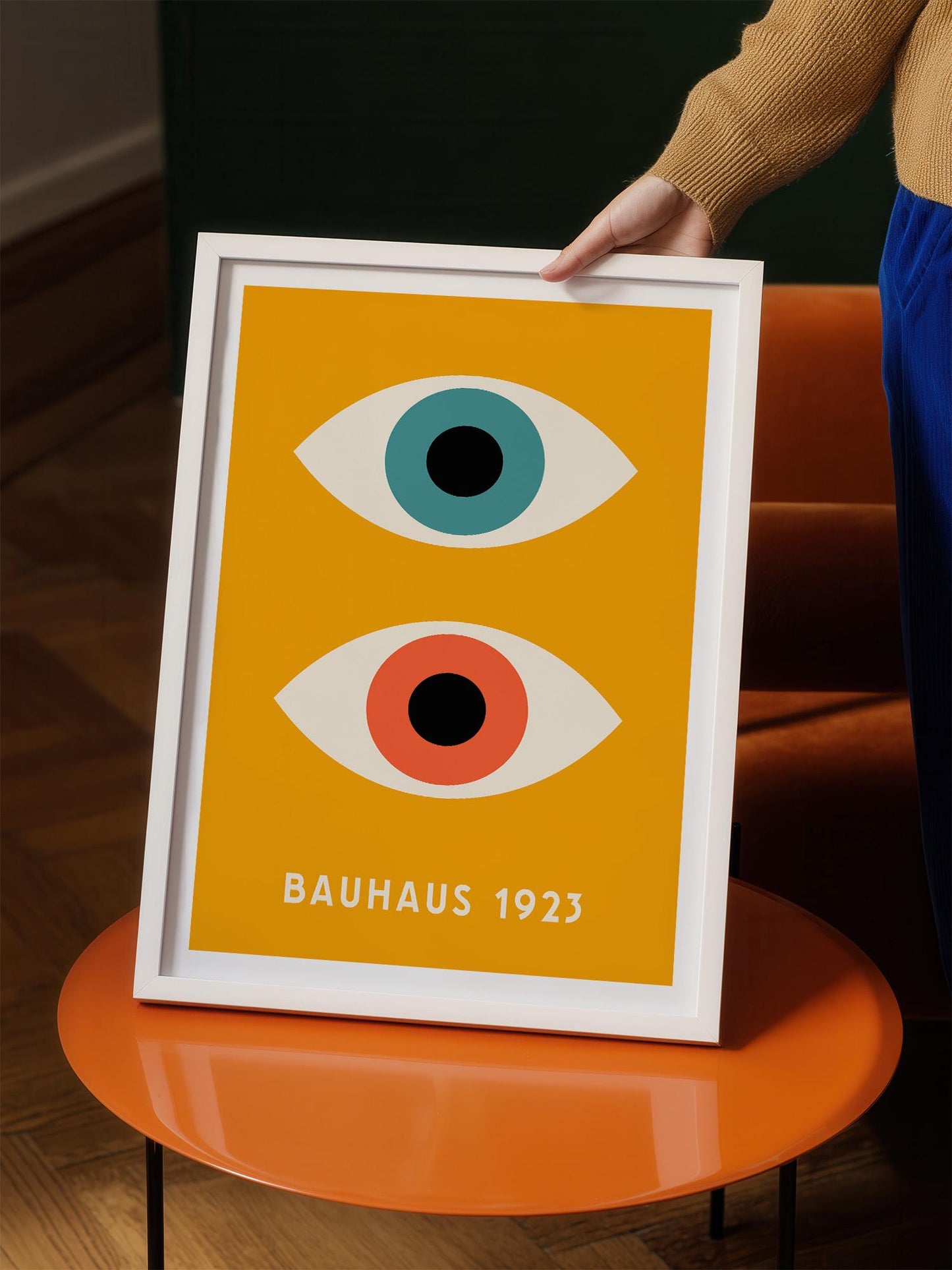 A person is holding up a framed poster with a yellow background and two stylized eye designs.